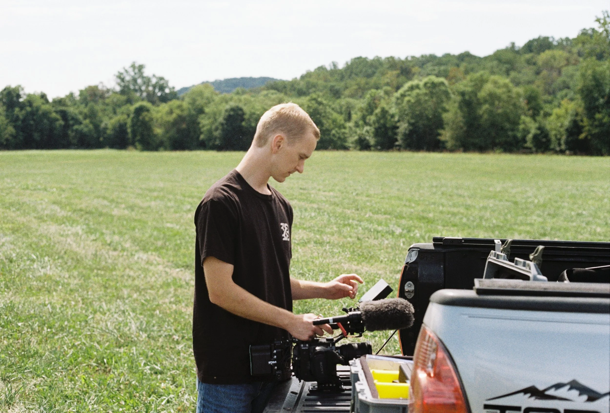 Young man standing in a field with a camera, setting up for filming, with trees and hills in the background.