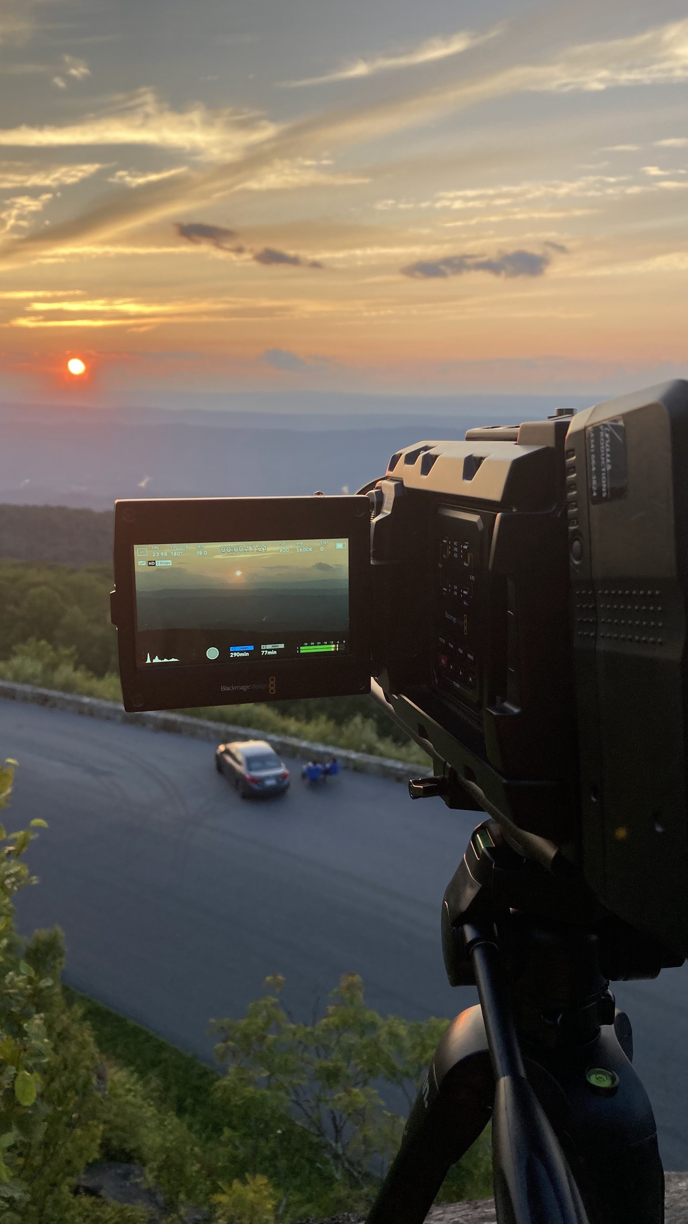 Professional camera on tripod capturing a sunset over a scenic landscape with a road, car, and a distant horizon.