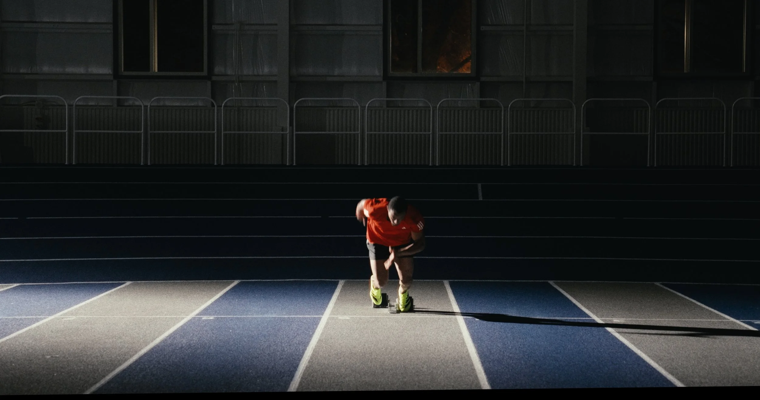 A male athlete in a red shirt and yellow running shoes bent over on an indoor track, preparing to race.