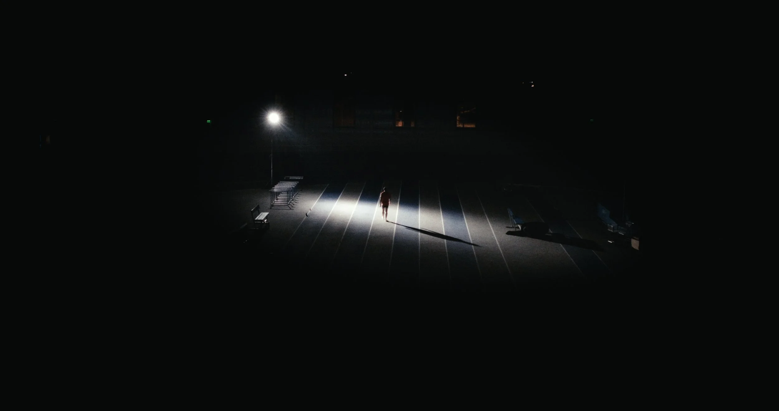 A person standing alone in a mostly empty parking lot at night, illuminated by a bright streetlight, with vacant benches and a few parked cars in the background.
