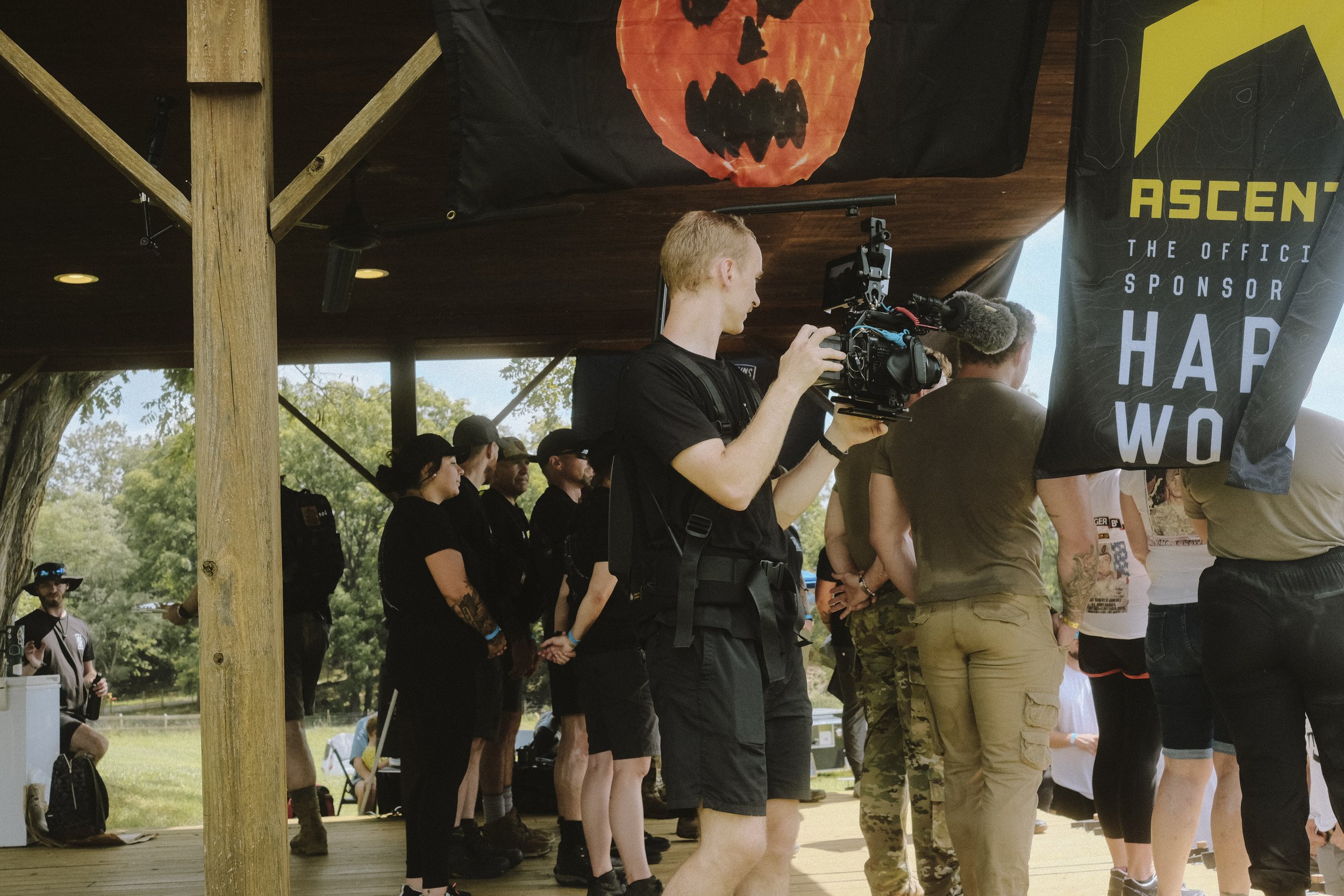 A group of people gathered on a wooden platform, with one person operating a professional video camera. A black flag with a jack-o'-lantern face and another flag with text hang above. The setting appears casual and outdoor, with trees visible in the