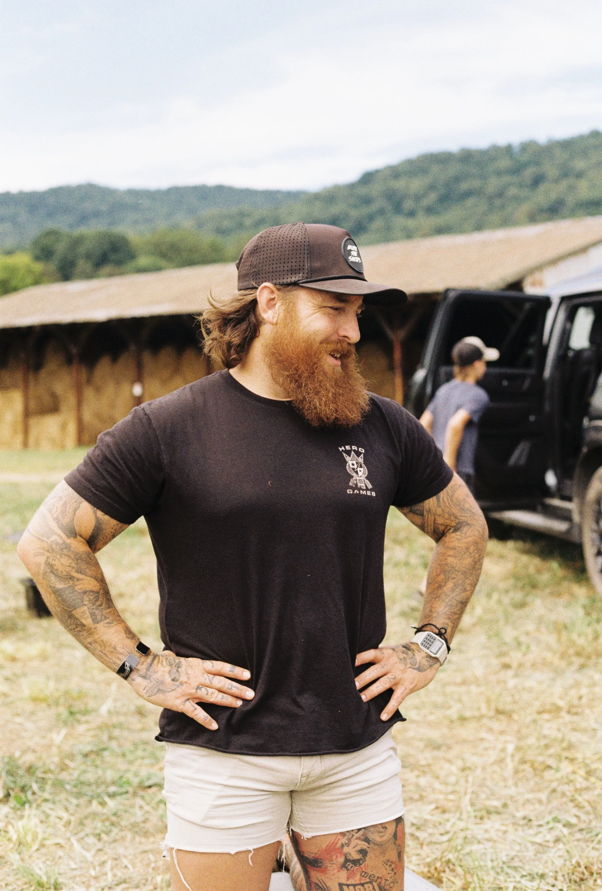 A bearded man in a black t-shirt and shorts standing outdoors with his hands on his hips, smiling. A woman in the background near a black vehicle, with a green mountain landscape behind them.