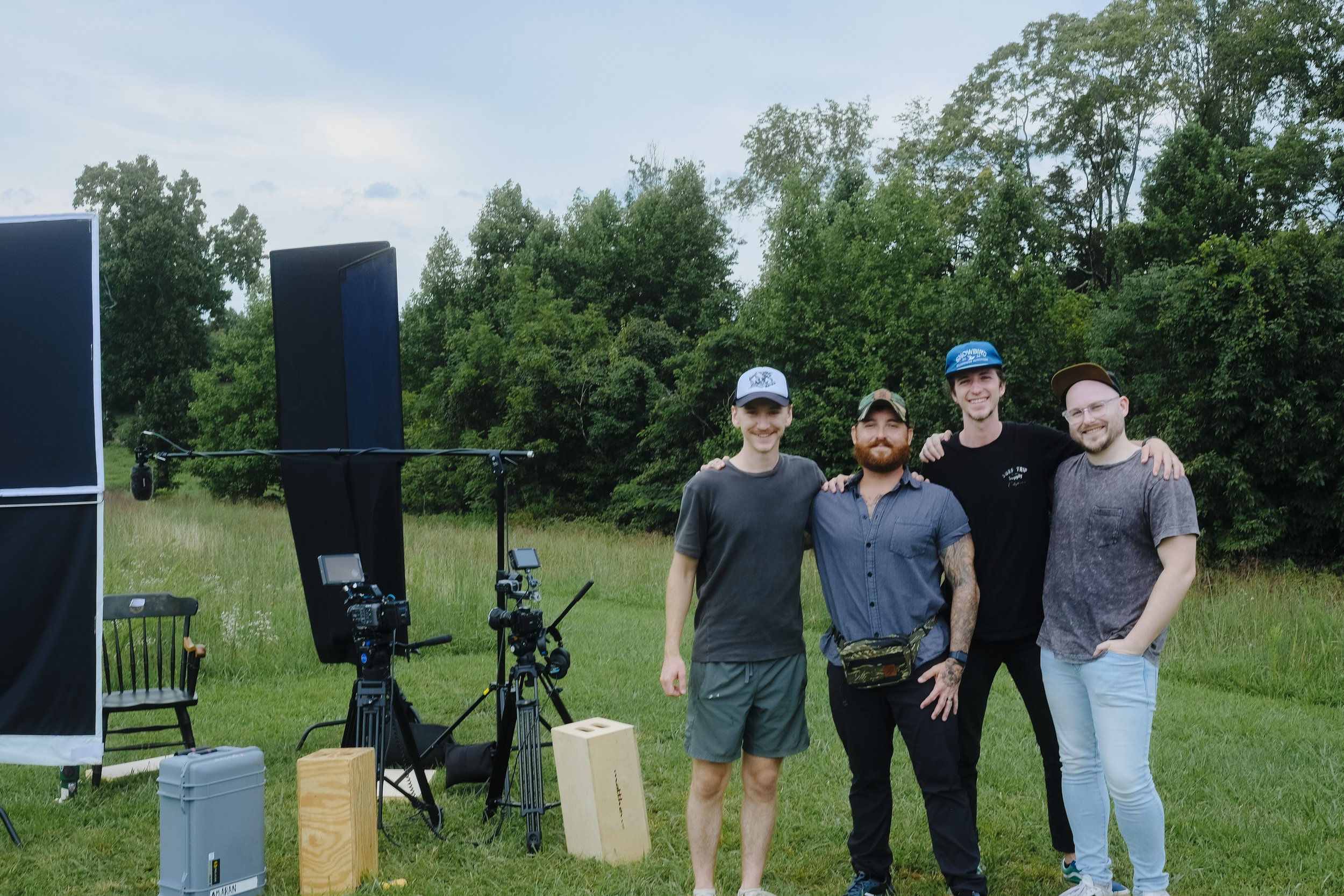 Four men standing together in a grassy field with film equipment around, including cameras on tripods and sound gear, surrounded by trees and bright sky.