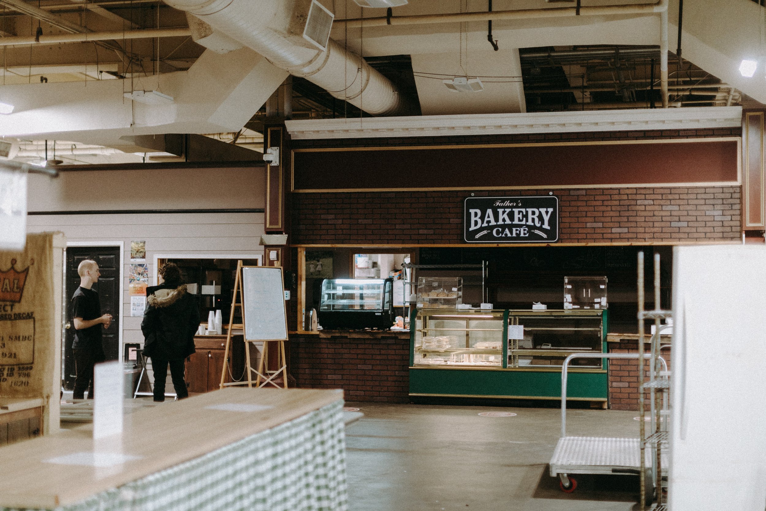 Interior of a bakery café with display cases and a café counter, two people standing nearby, and a sign reading "Bakery Café."