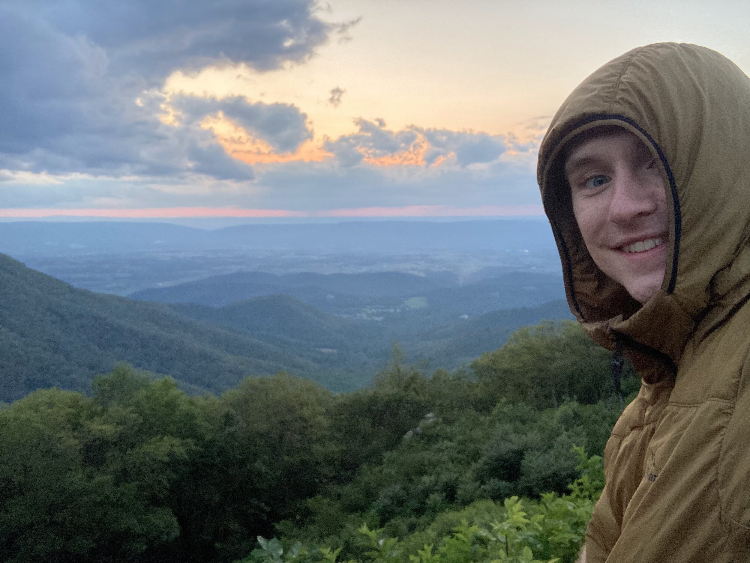 Smiling person in a brown hooded jacket standing on a mountain with a scenic view of hills and a colorful sky at sunset in the background.
