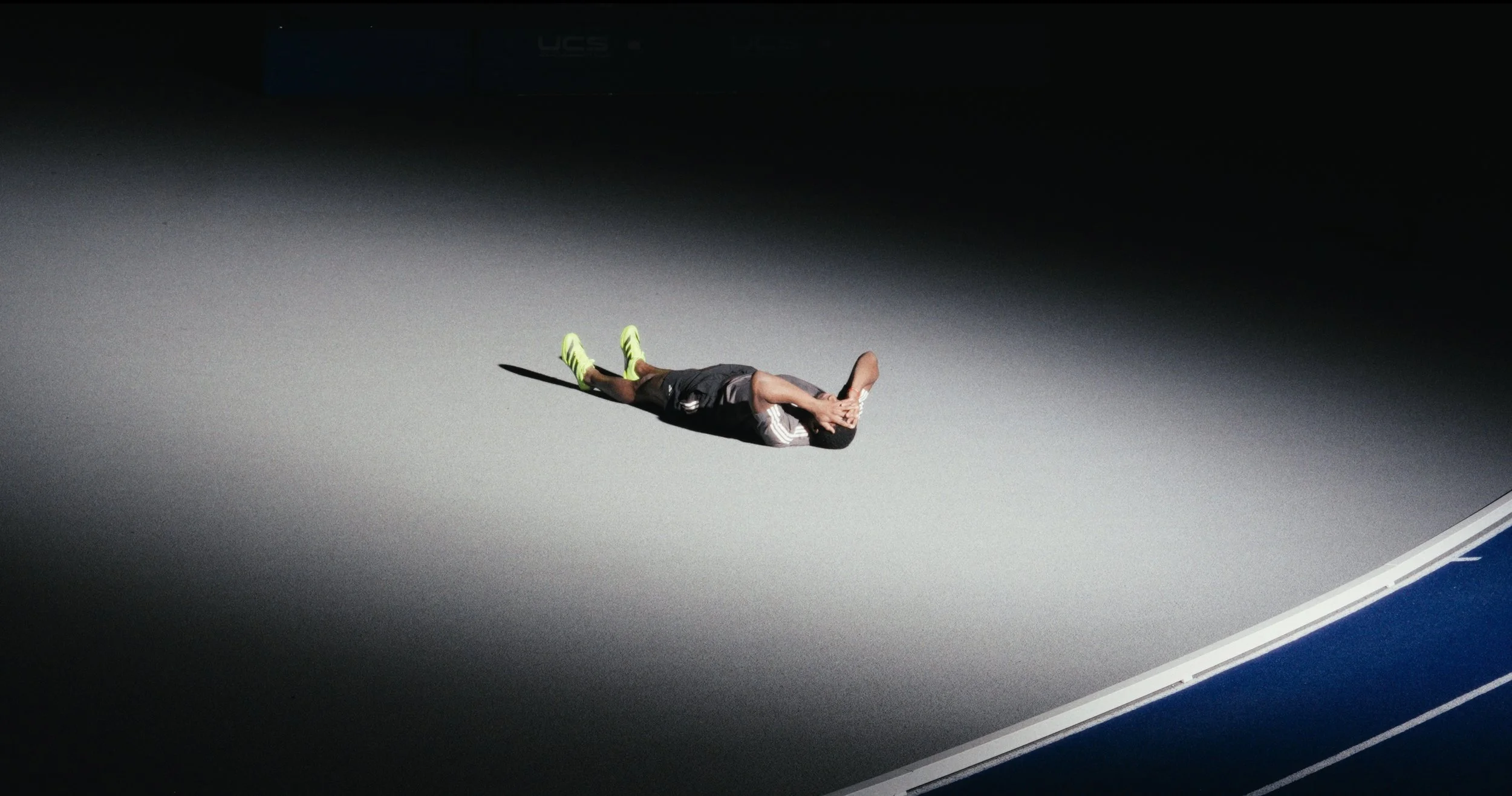 A tennis player lies on his back on a tennis court with his hands covering his face, illuminated by a spotlight amid darkness.