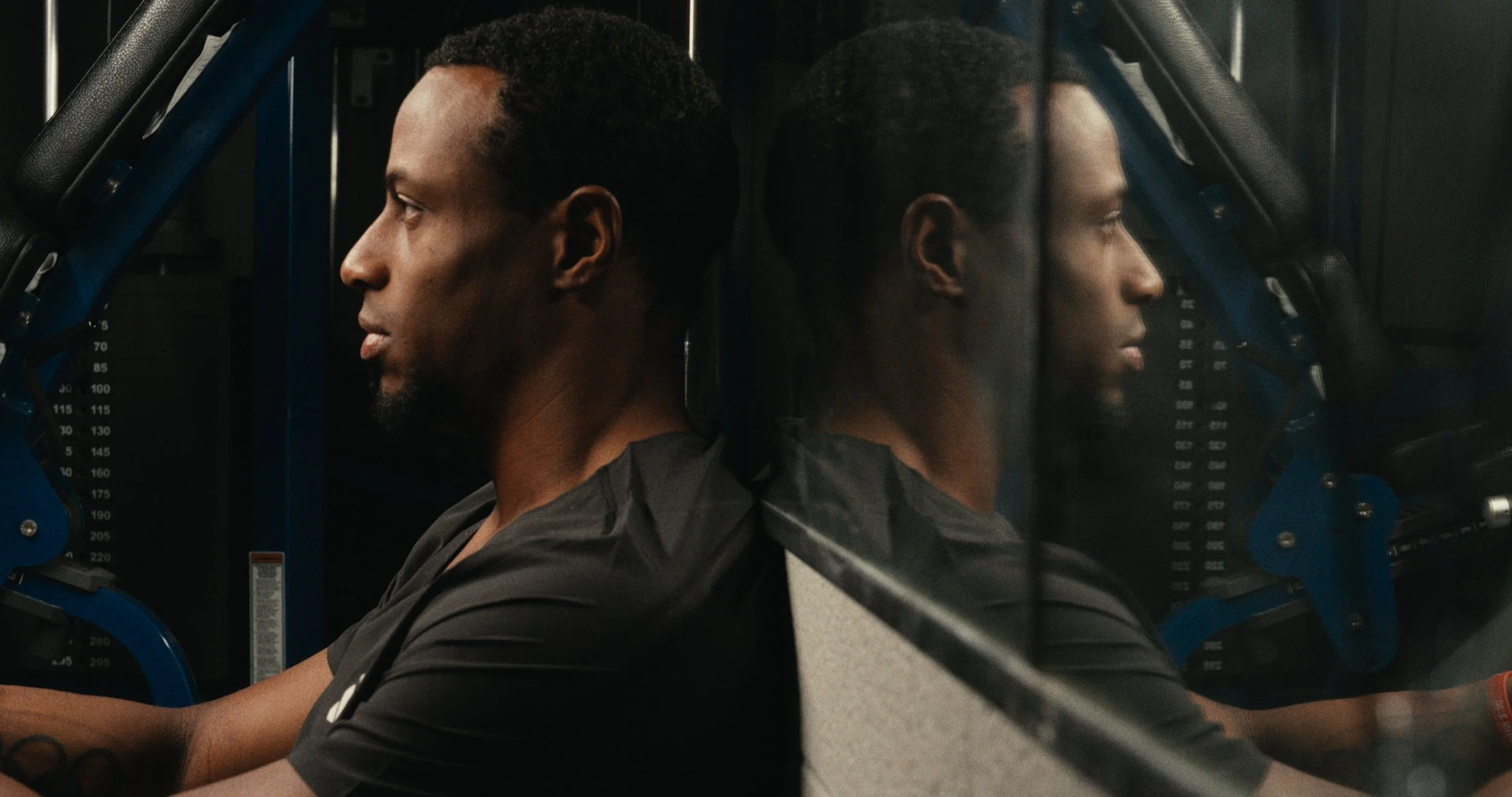 A man with a dark complexion, short curly hair, and a goatee is sitting indoors, looking to the left, with his reflection visible in a mirror.”}