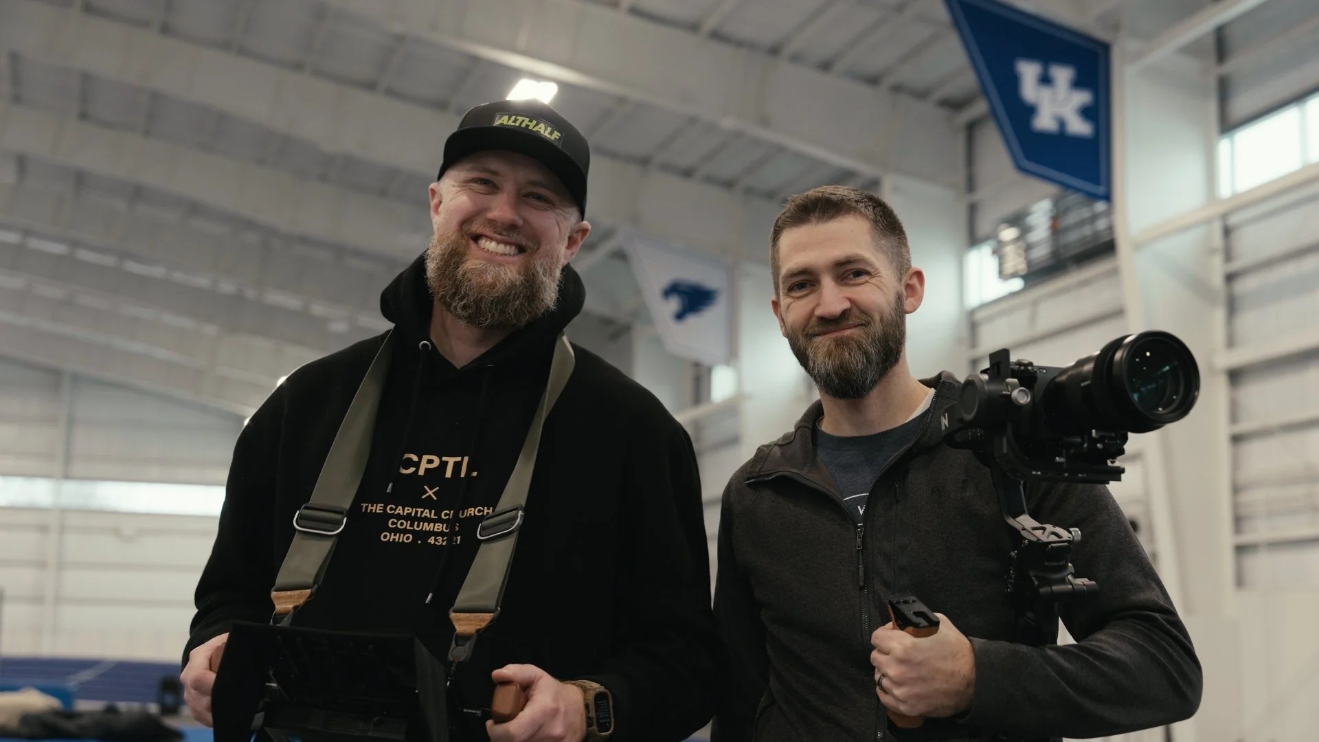Two smiling men with beards standing inside a sports facility, one holding a camera with a large lens and the other wearing a cap and a camera strap around his neck.