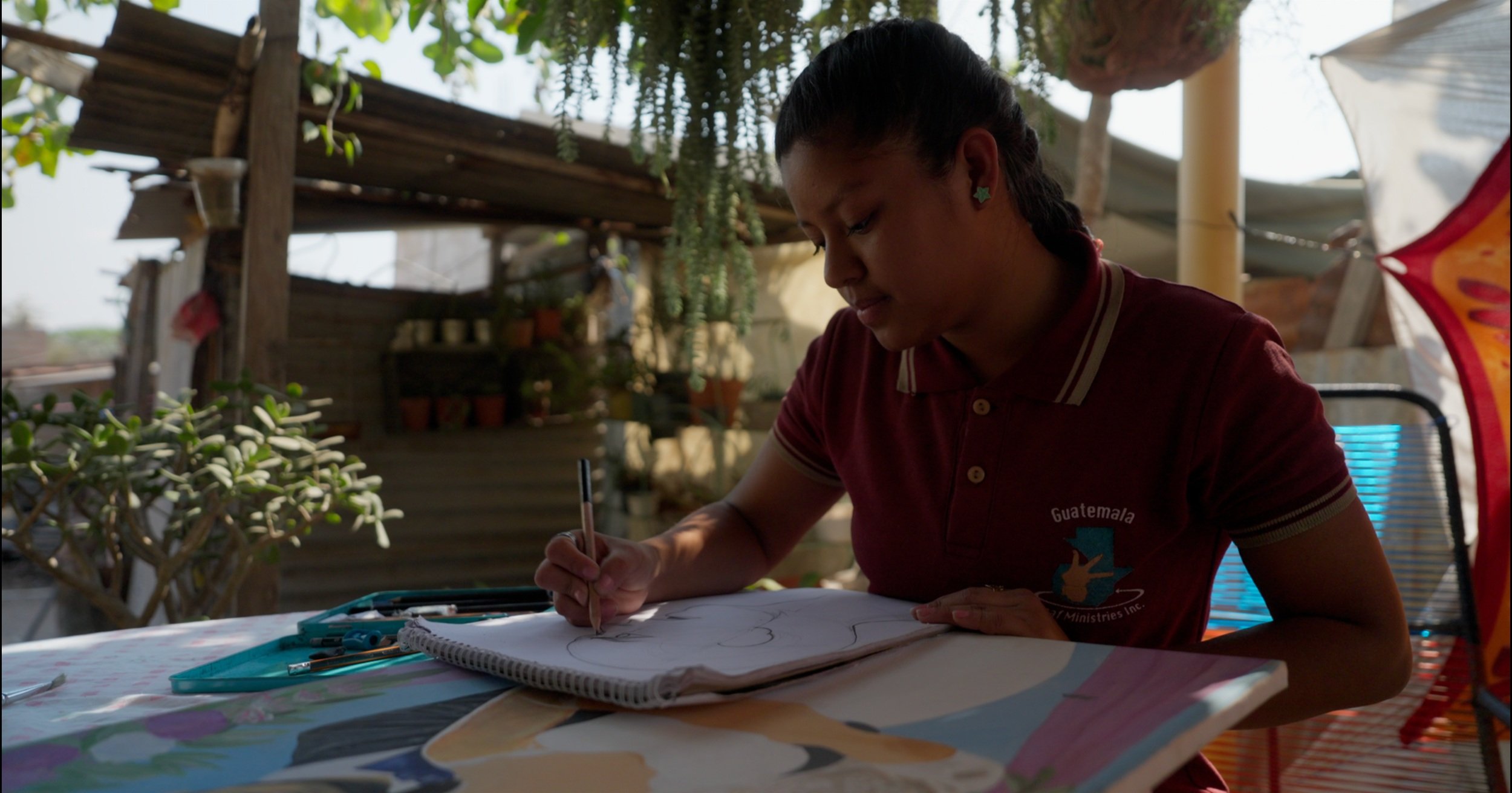 A young girl sitting at a table outdoors, drawing in a notebook with a pen under a shaded area with plants and wooden structures.