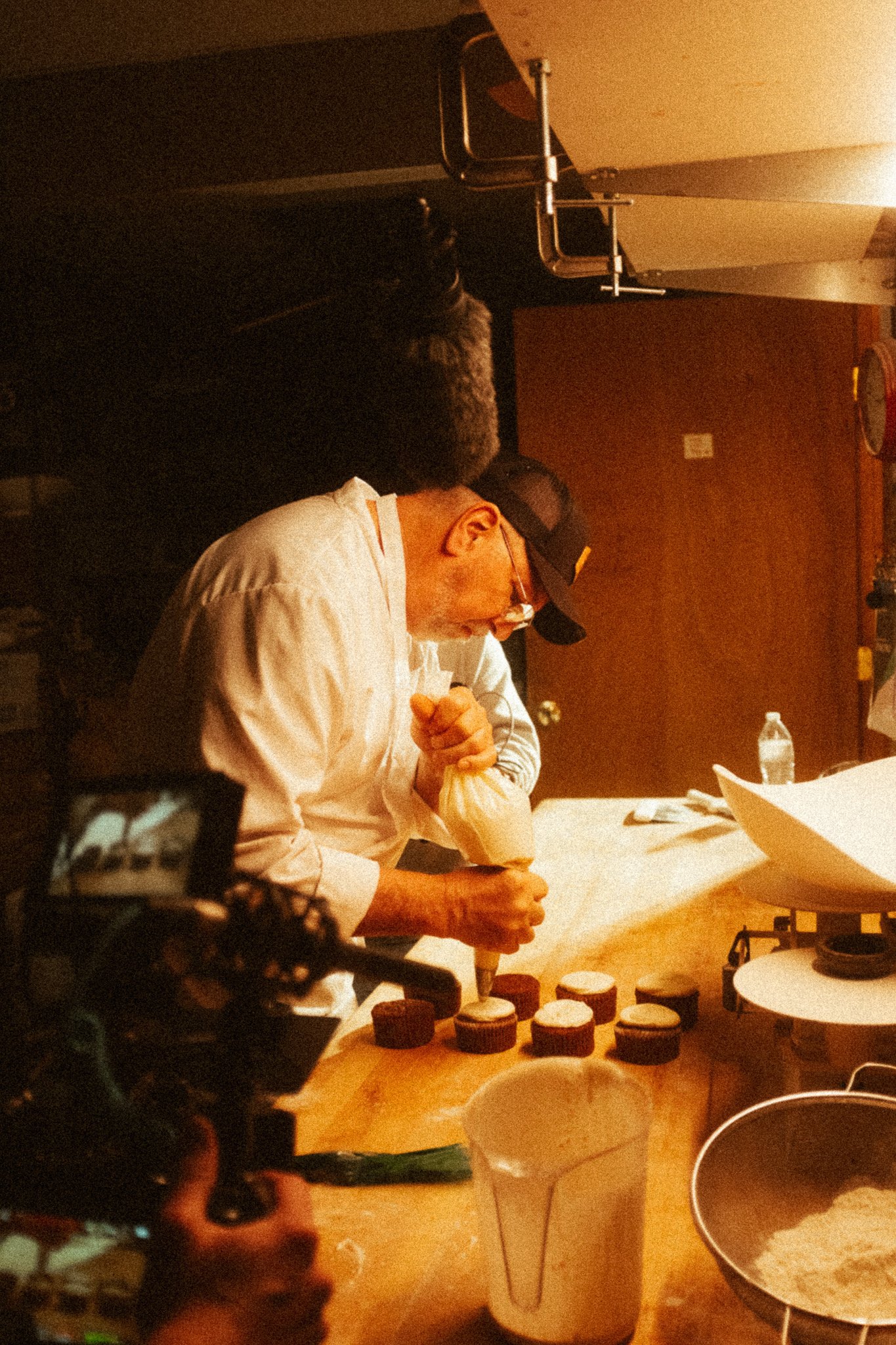A chef in a kitchen piping frosting onto cupcakes.