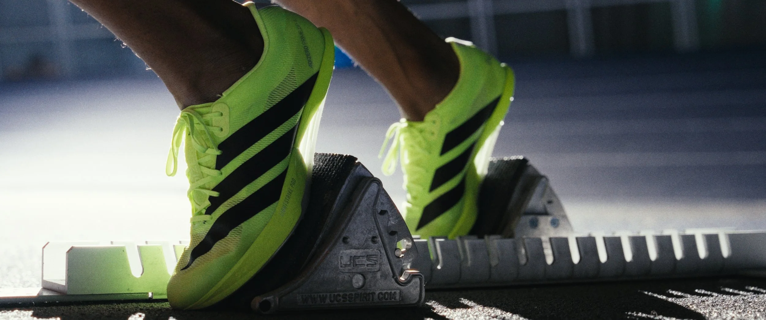 Close-up of a person's feet wearing bright yellow running shoes with black stripes, positioned on a starting block at a track, ready to race.