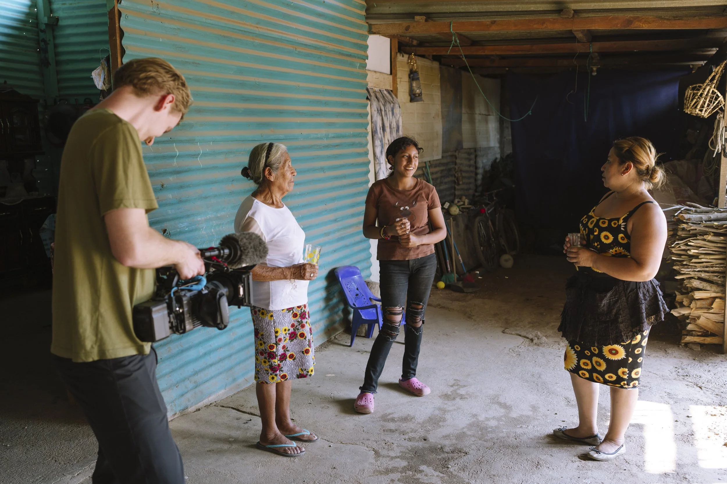 A woman being interviewed by a camera crew in a rural, semi-outdoor space with corrugated metal wall, wooden ceiling, and stacked wood in the background in Guatemala.