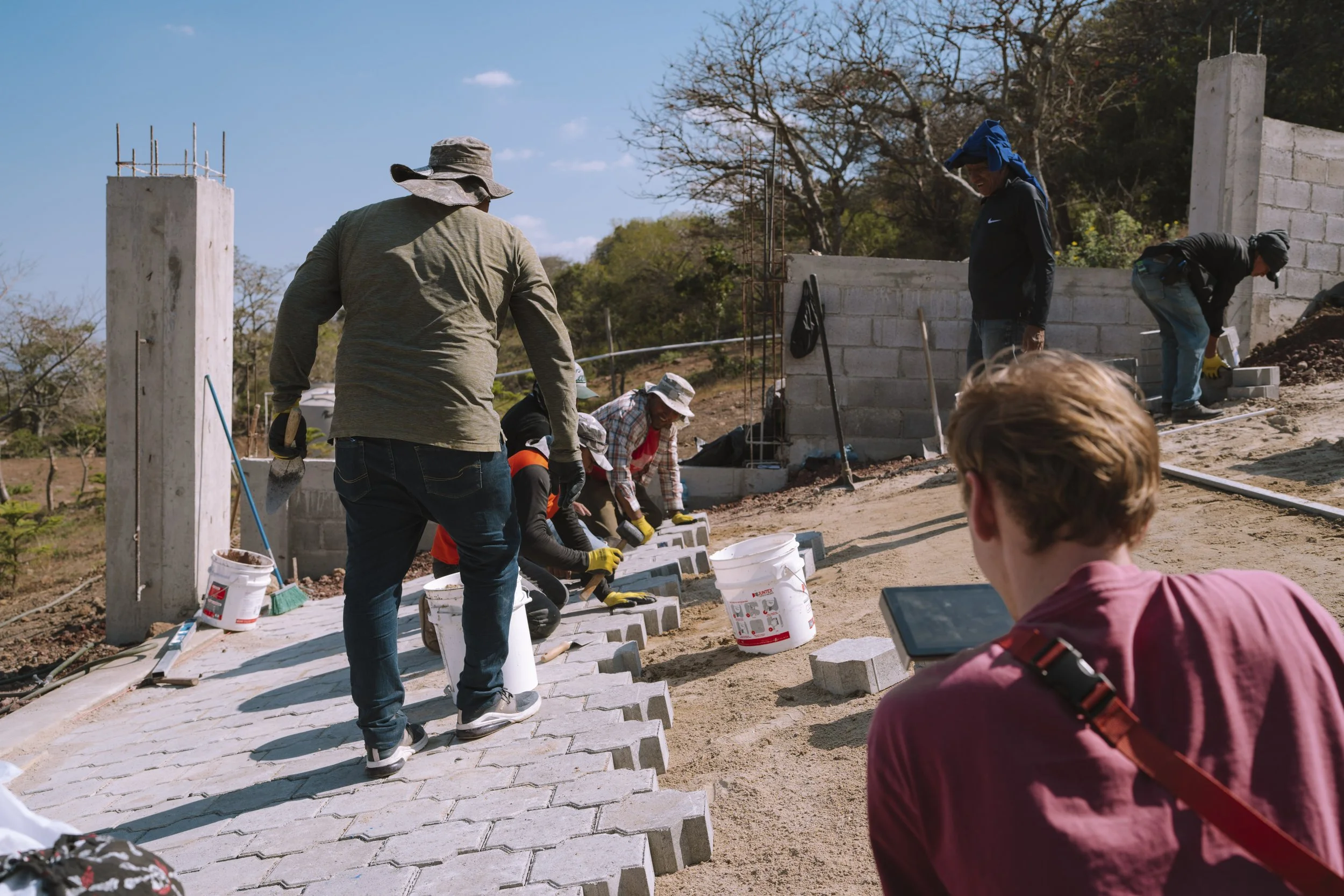 Group of people working together to build an outdoor driveway for the school with interlocking pavers, some kneeling and some standing, with construction tools and buckets around.