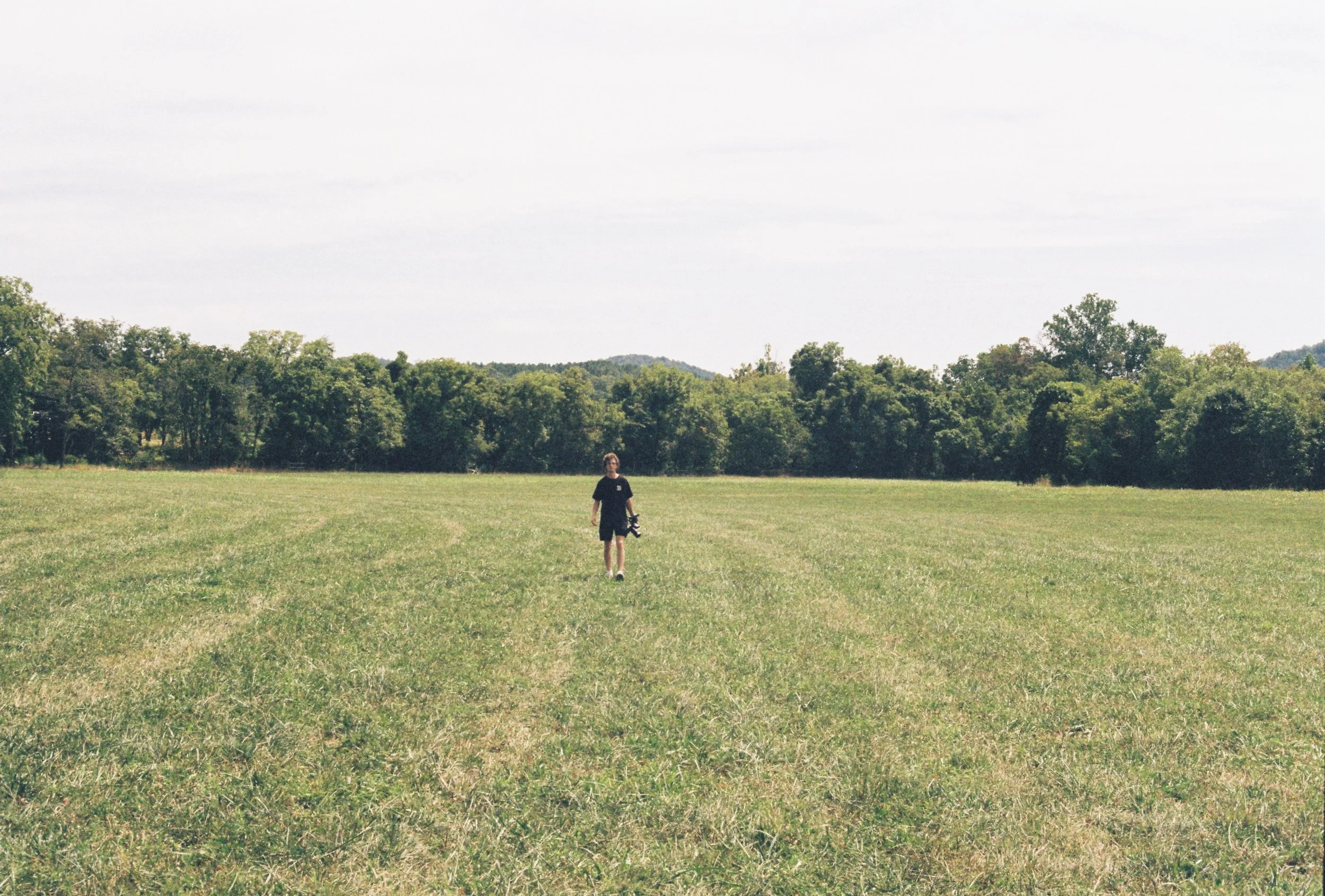 Person walking across a grassy field with trees and hills in the background on a bright, clear day.