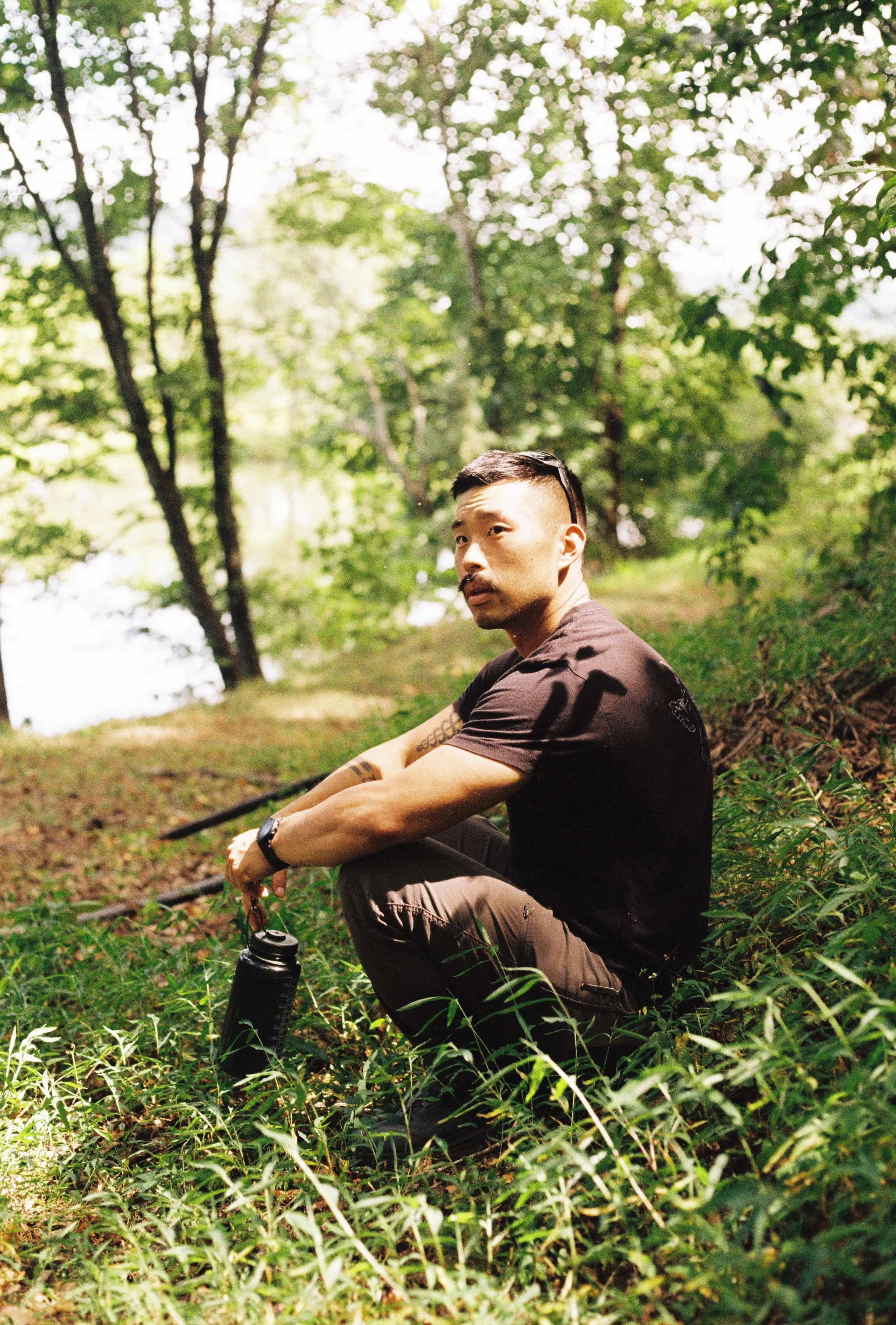 A man sitting cross-legged outdoors in a forested area, holding a water bottle, with trees and greenery in the background.