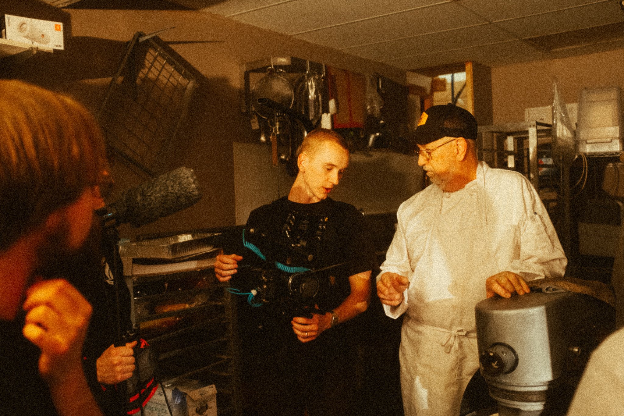 A group of people, including a filmmaker with a camera rig and a man in a chef's coat, talking in a commercial kitchen.