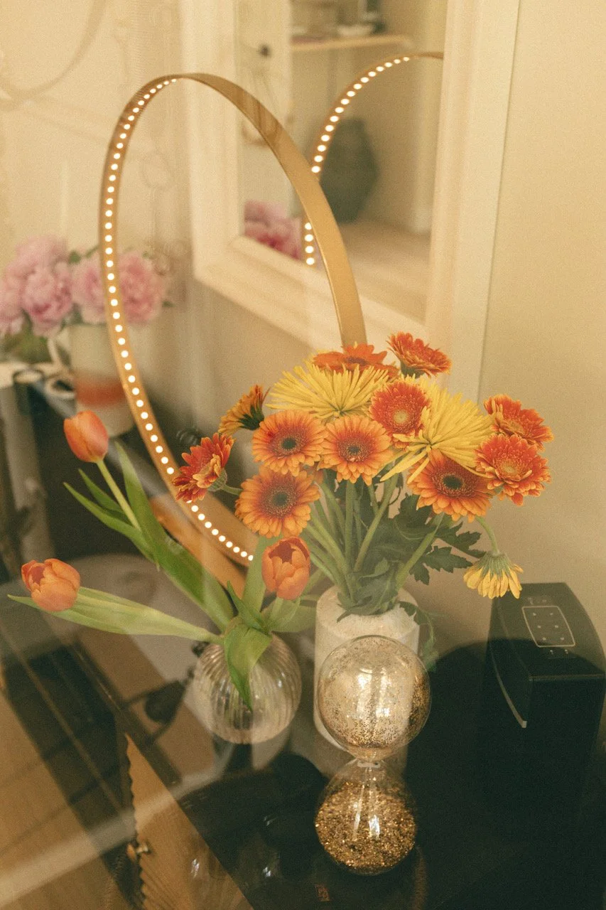 Miroir en forme de cœur avec lampes LED, vase avec fleurs orange et jaunes, miroir à fond blanc, objets décoratifs dorés sur une table en verre.