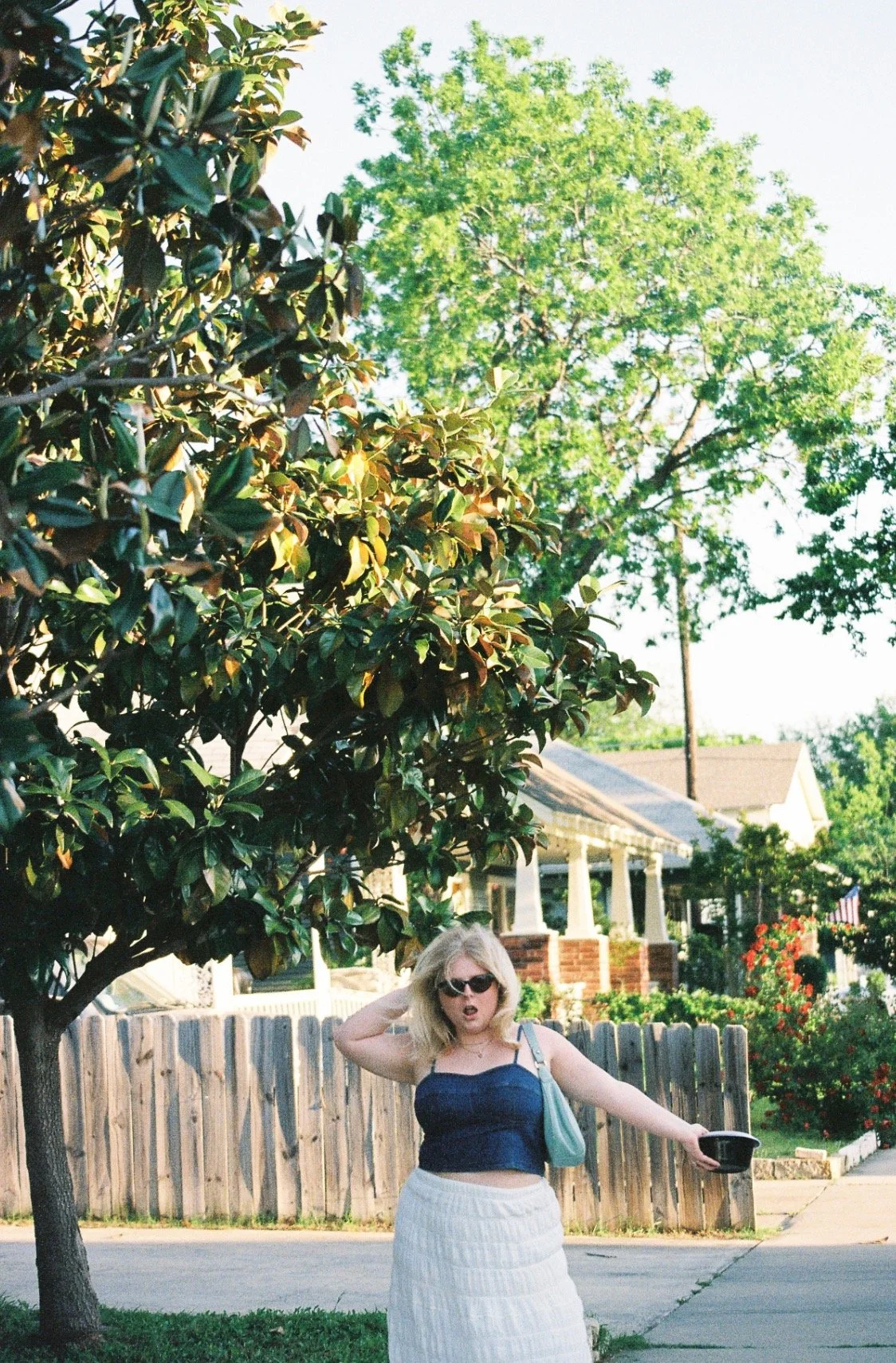 Woman with sunglasses and a blue top standing near a tree on a residential street, holding a black container.