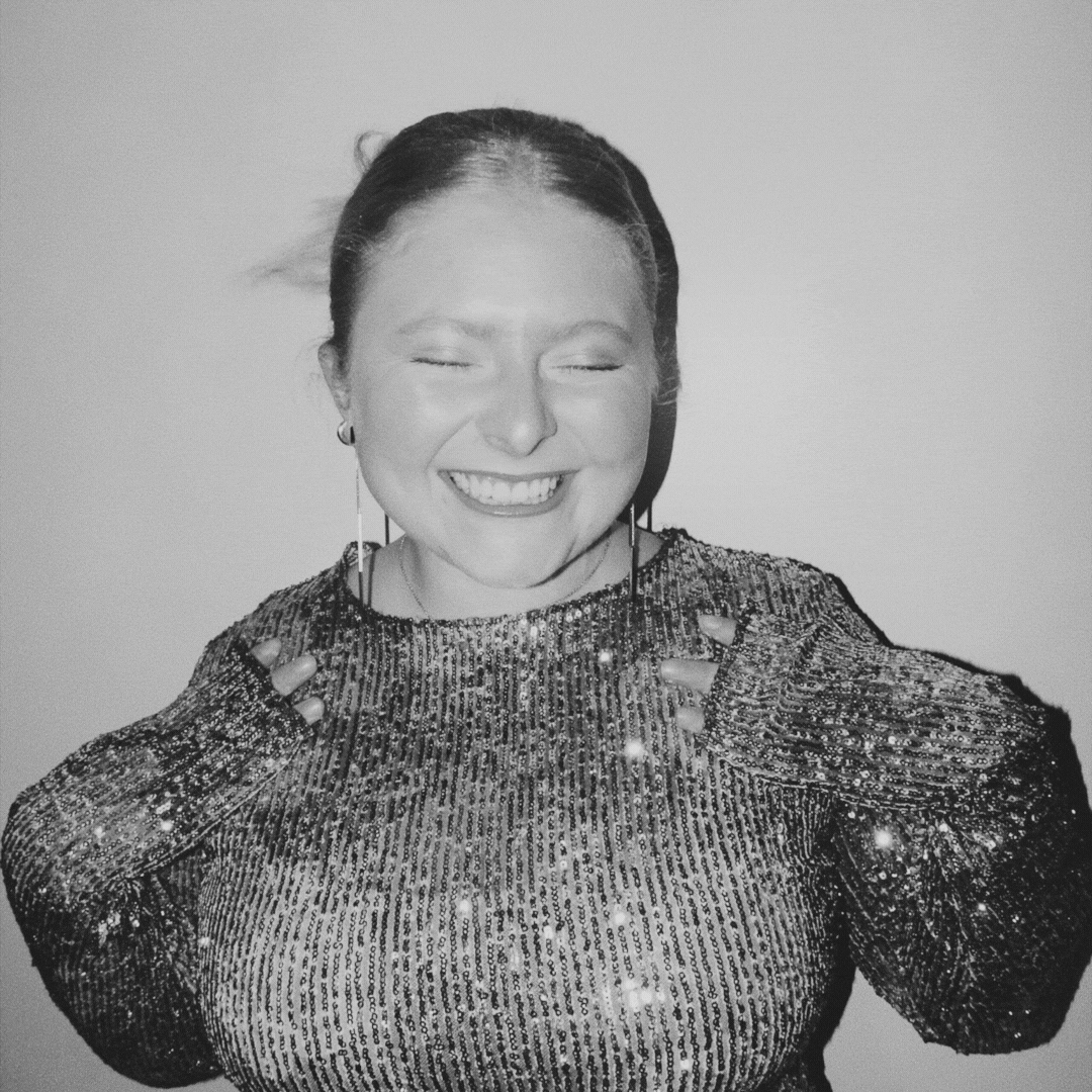 Black and white photo of a person smiling with eyes closed, wearing a sparkly sequined top and earrings.