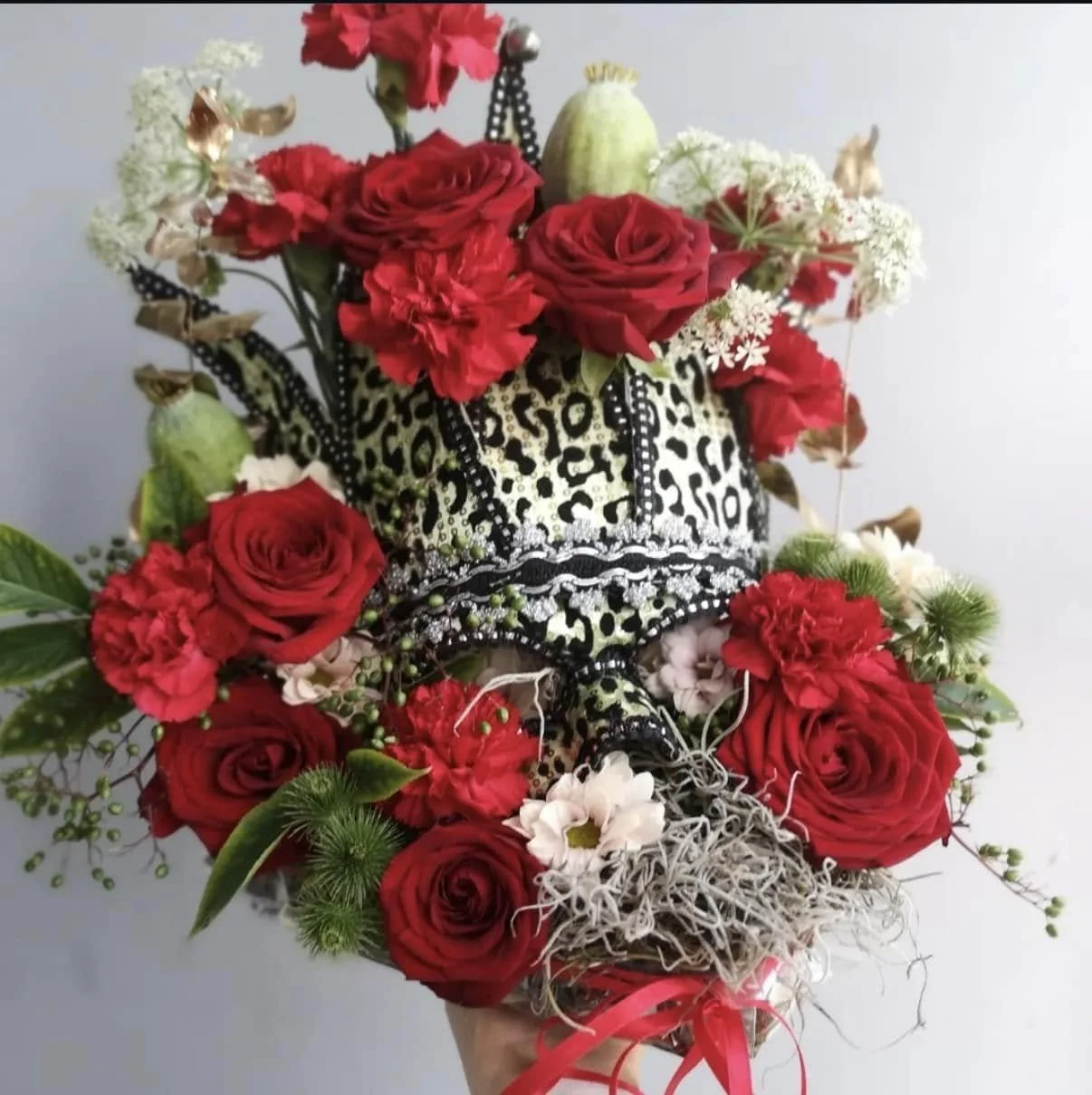 A floral arrangement featuring red roses, red carnations, white flowers, and greenery, arranged around a leopard-print crown with black and silver embellishments, tied with a pink ribbon.
