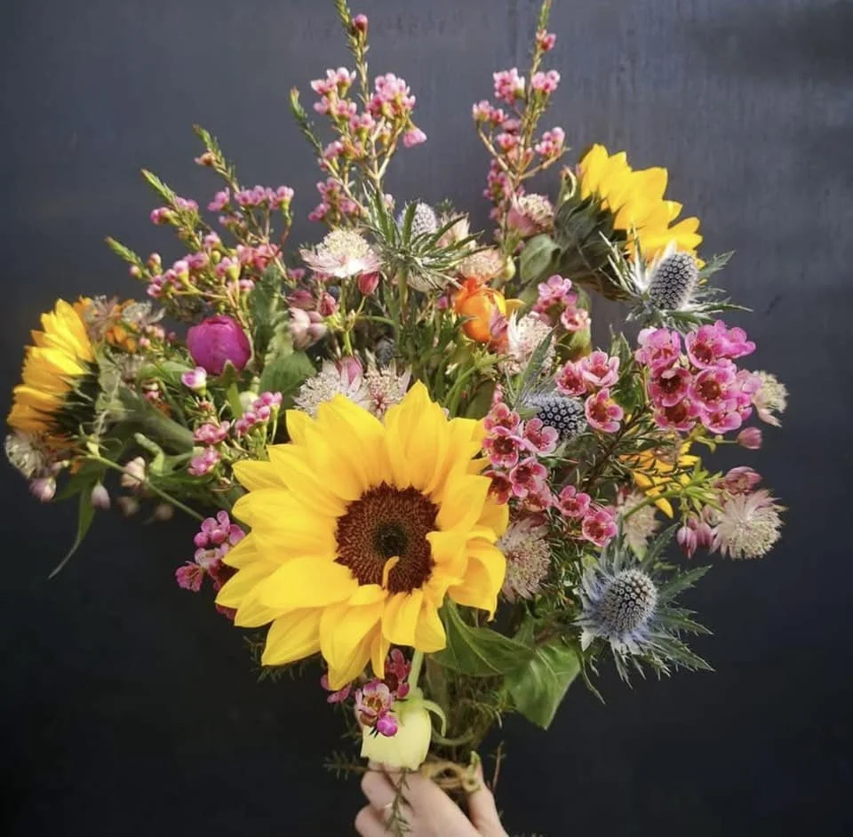 A colorful bouquet of flowers featuring a large sunflower, pink wax flowers, white and purple thistle, and small white and pink blossoms, held by a person against a dark background.