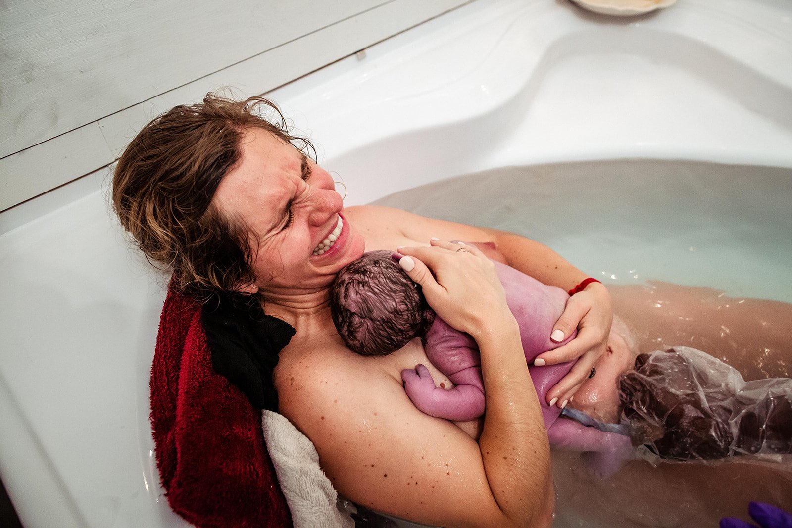 A woman is smiling and holding a newborn baby in a birthing tub during a water birth.