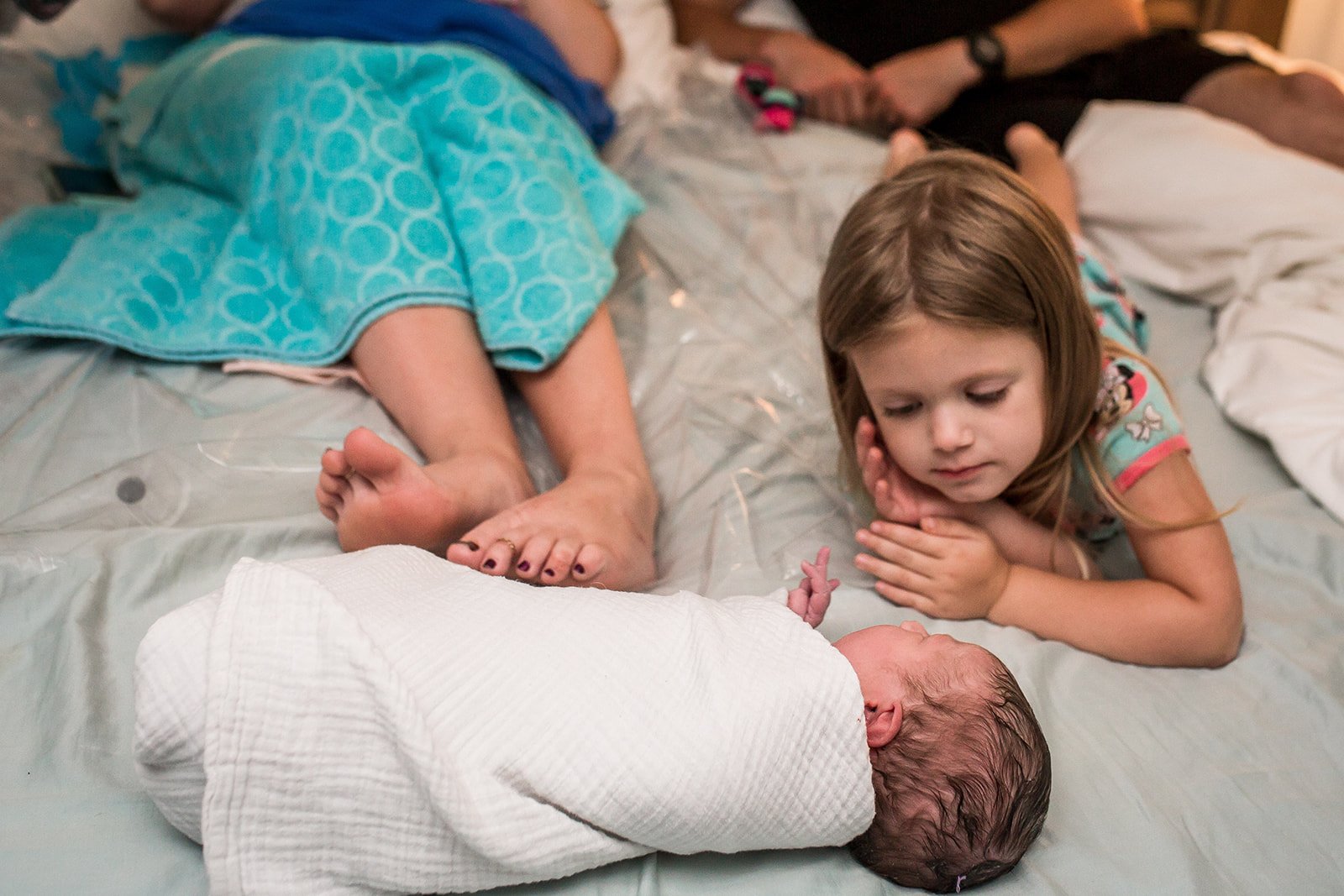 Young girl lying on a bed looking at a newborn baby wrapped in a white blanket.