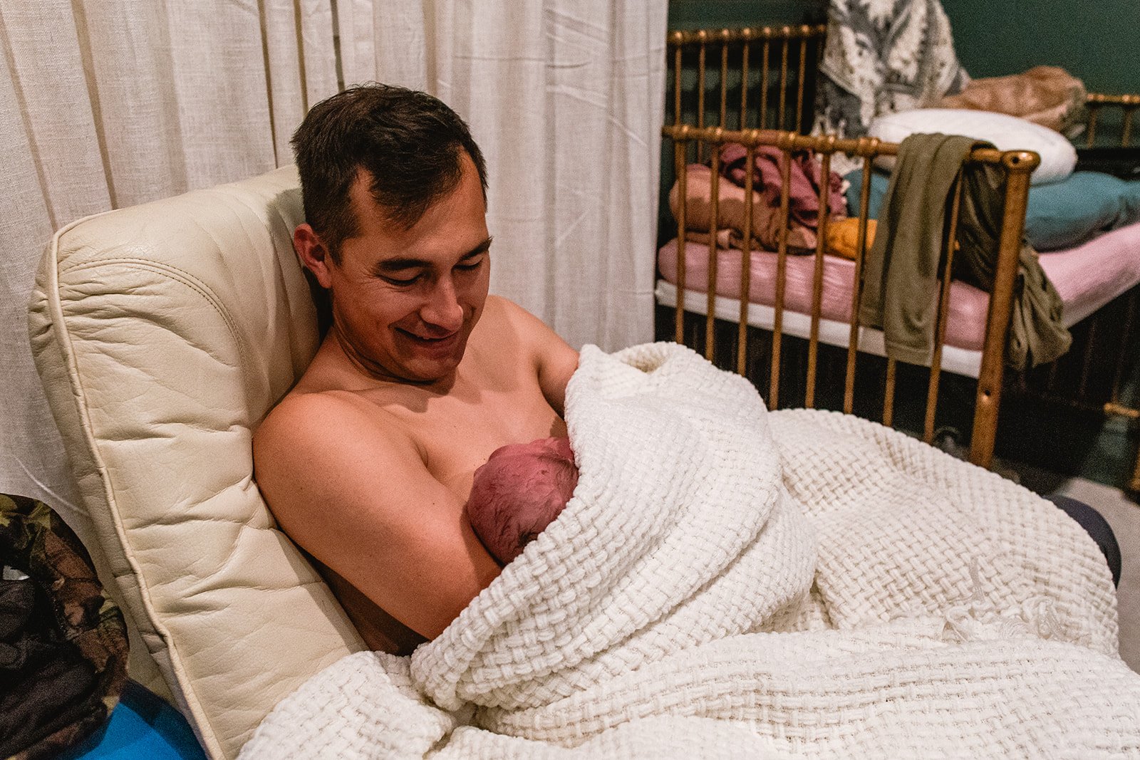 Man holding newborn baby wrapped in a white blanket, sitting on a chair next to a crib. Home birth, water birth, prenatal and postpartum midwifery care, Pooler, GA, Savannah, GA Midwife