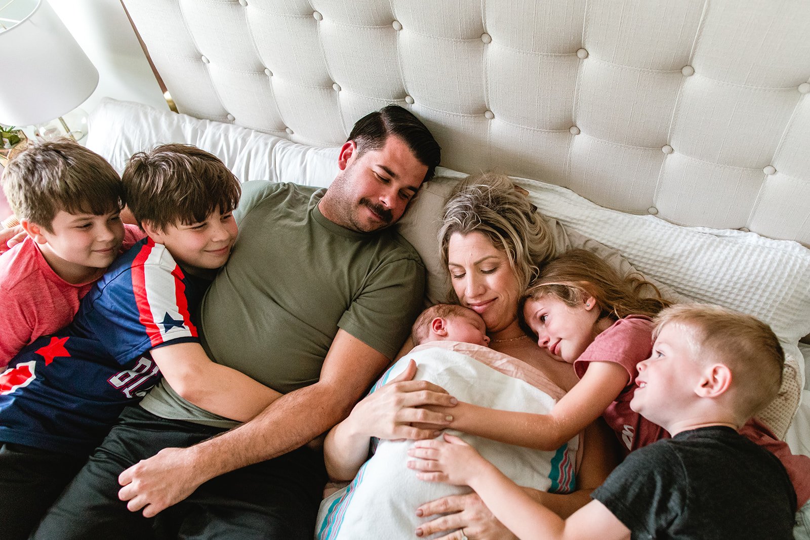 A family of six lying on a bed, with the parents cuddling a newborn baby, displaying expressions of happiness and affection, in a cozy bedroom setting. Home birth, water birth, prenatal and postpartum midwifery care, Pooler, GA, Savannah, GA Midwife