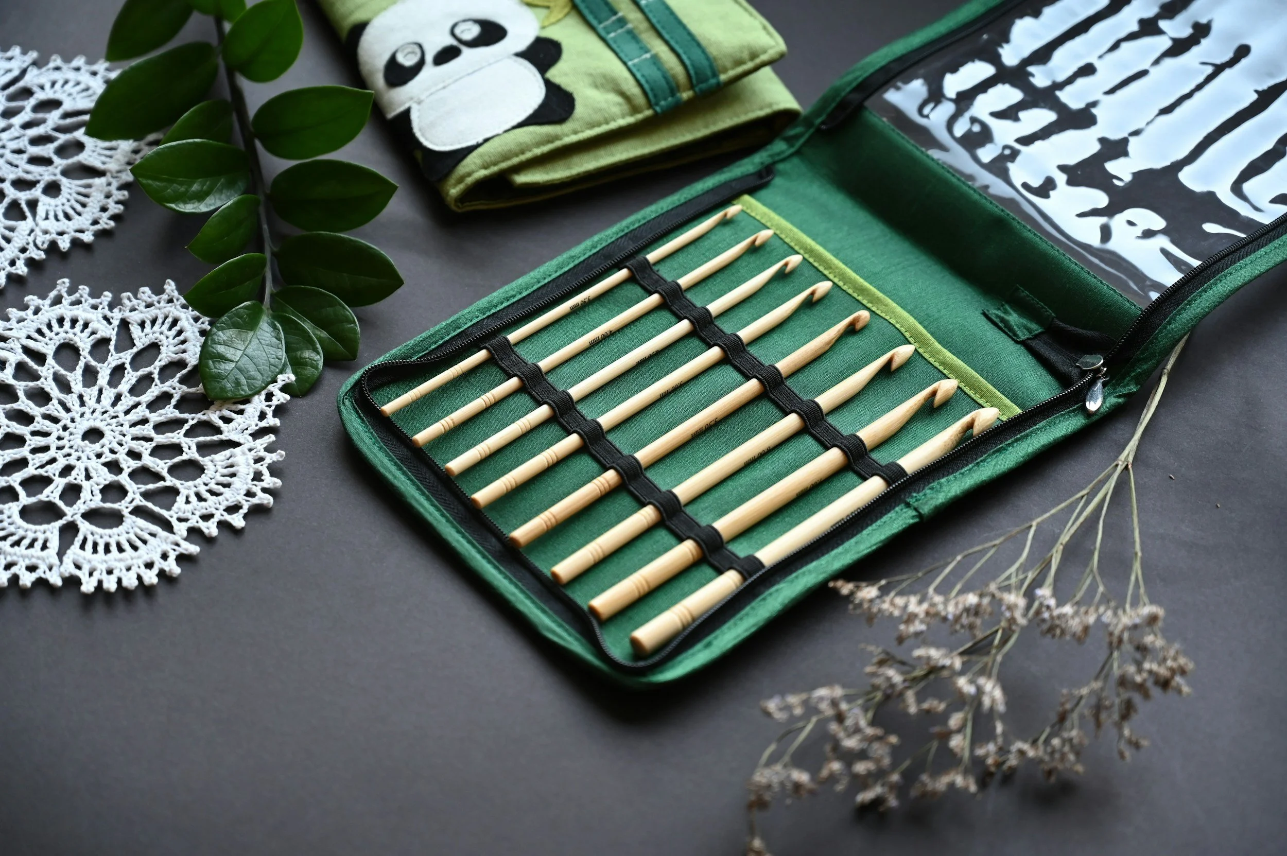 Set of wooden crochet hooks in a green case, surrounded by crochet doilies, a green leafy branch, a fabric item with a panda face, and dried flowers on a dark surface.