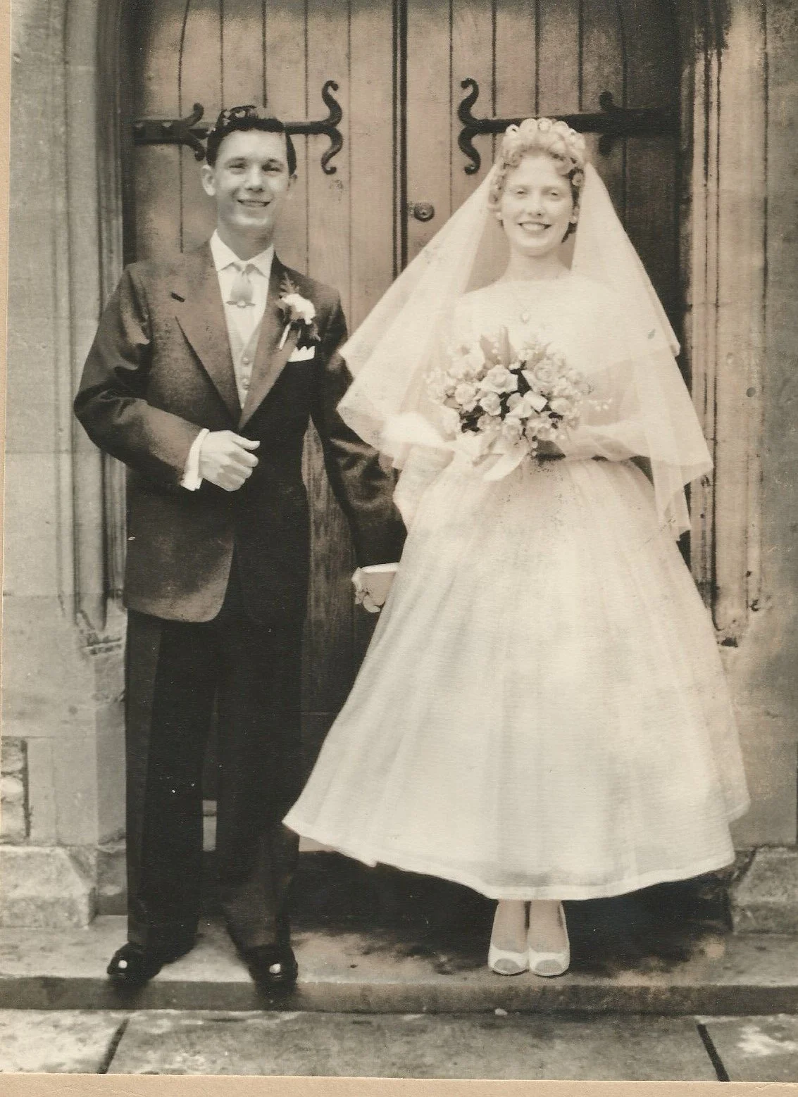A black and white photograph of a bride and groom standing in front of a wooden door with decorative black wrought iron hinges. The groom is dressed in a dark suit with a boutonnière, and the bride is wearing a wedding dress with a full skirt, veil, and holding a bouquet of flowers.