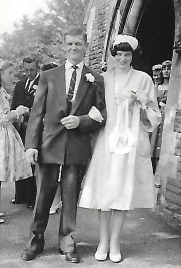 A black and white photo of a man and woman, likely at a wedding, walking arm-in-arm outside a brick building. The man is wearing a suit with a tie and a boutonniere, while the woman is in a dress with a floral headpiece.