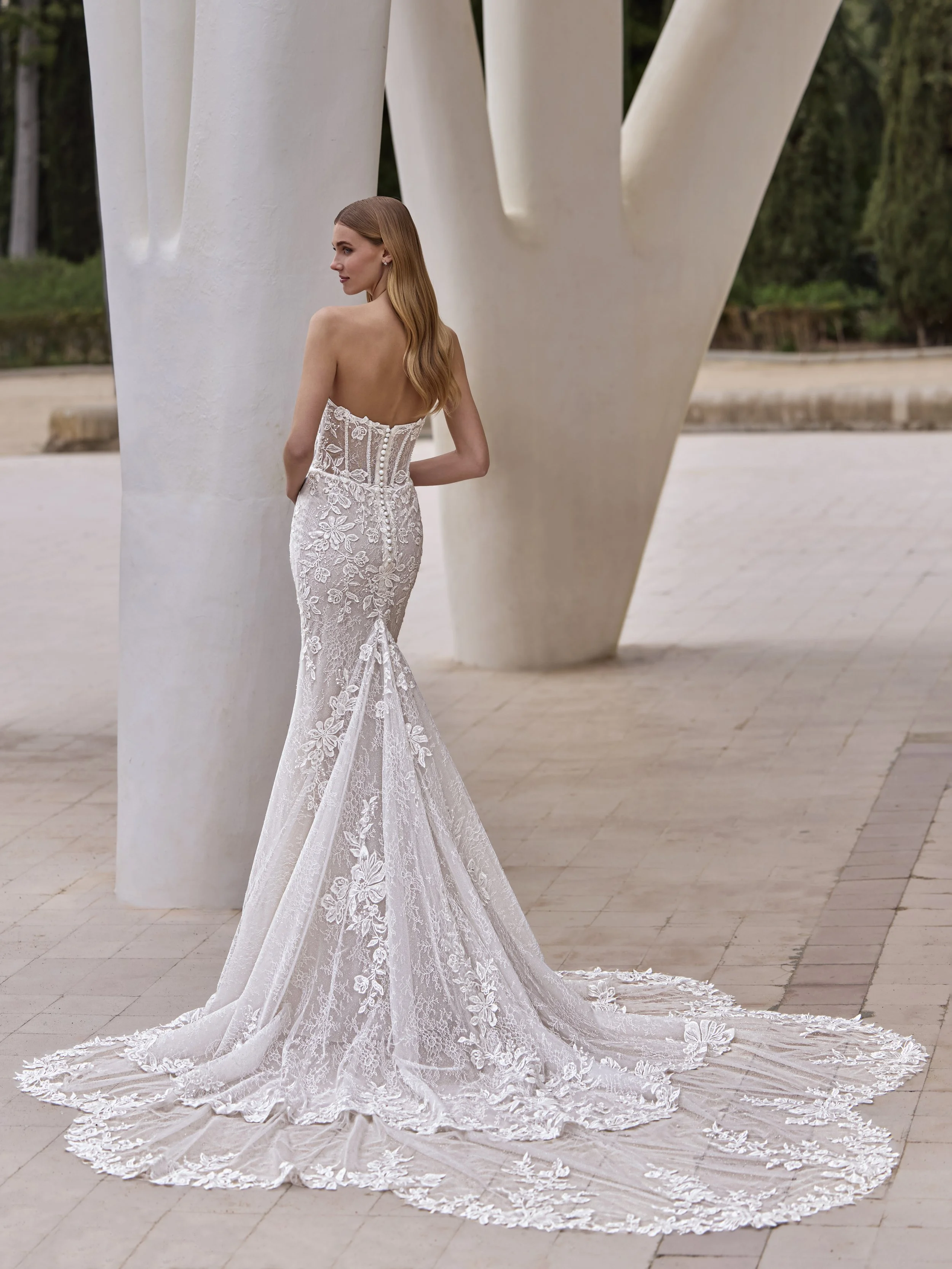 A woman in a white wedding dress with floral embroidery and a long train, standing outdoors near white modern sculpture structures.