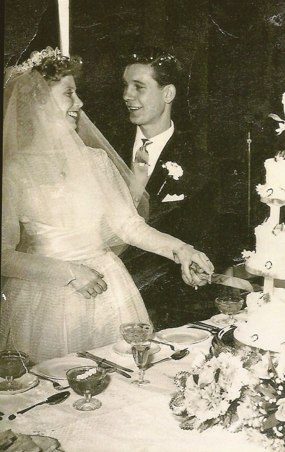 A black-and-white photo of a bride and groom cutting a wedding cake together, smiling at each other, at their wedding reception. The table has glasses, plates, and a floral centerpiece.