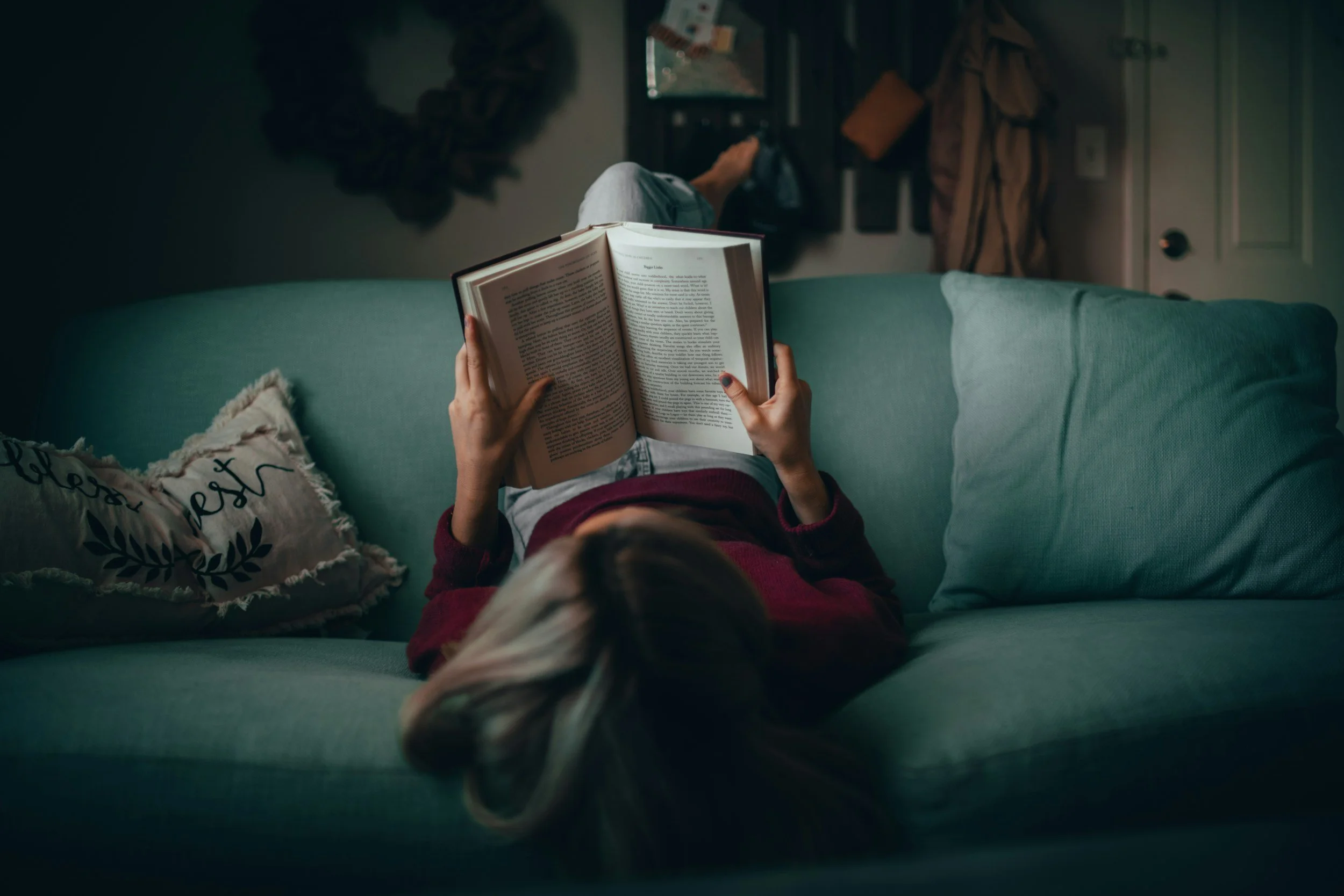 Person lying on a teal sofa, reading a large book, with a pillow that says 'love' on it, in a cozy living room with a mirror and coat hooks in the background.