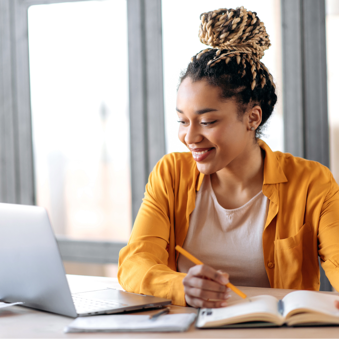 A young woman with braided hair in a bun, wearing a mustard-colored shirt, sitting at a desk with a laptop, open notebook, and pencil, smiling while studying in a well-lit room with large windows.