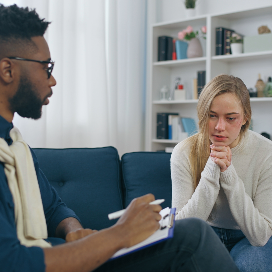 A woman with long blonde hair and a white sweater sits on a blue couch, looking distressed, as she talks to a man with glasses, a beard, and a dark shirt, who is taking notes on a clipboard in a room with a bookshelf in the background.