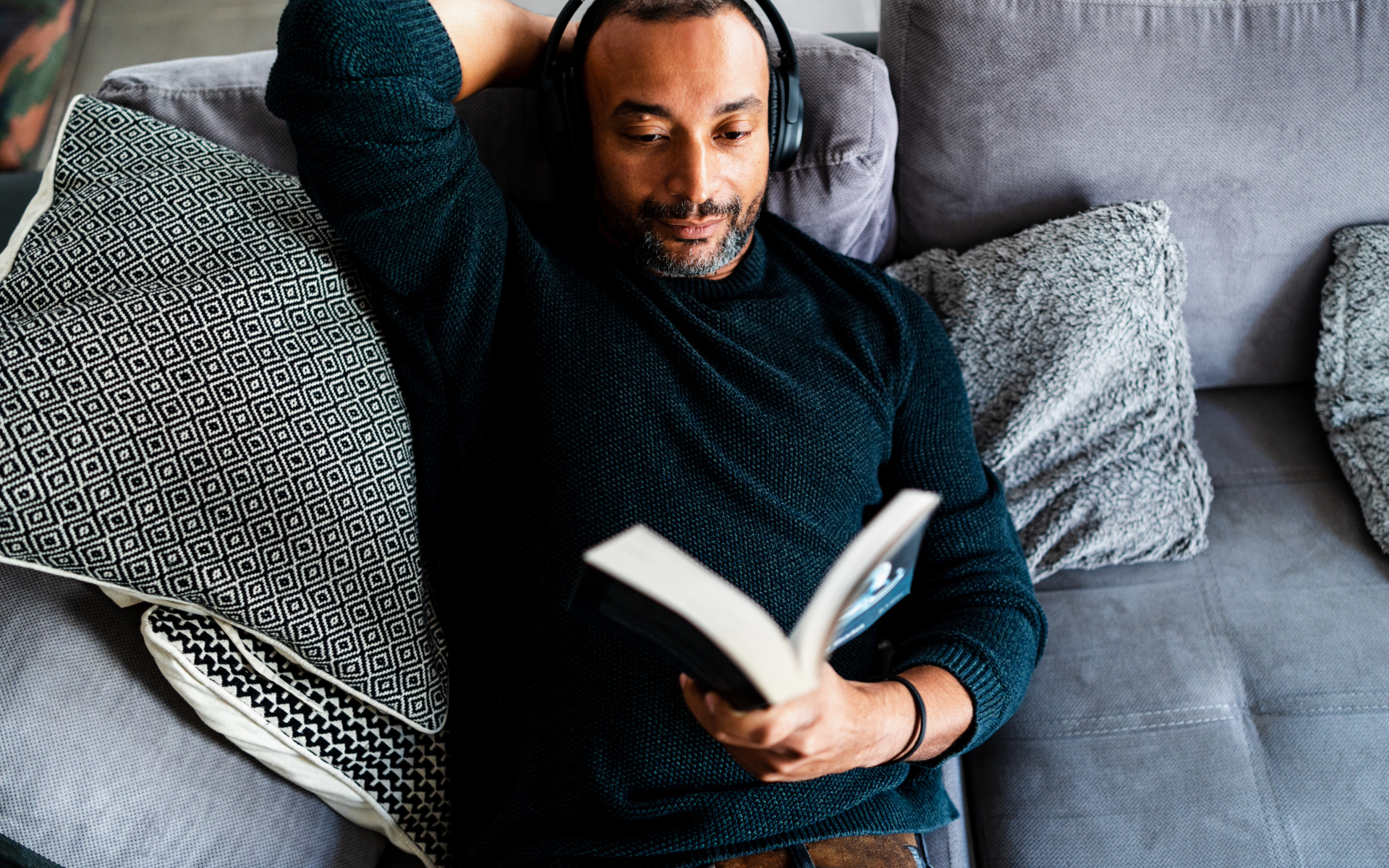 A man with headphones reading a book while lying on a gray sofa, surrounded by decorative pillows.