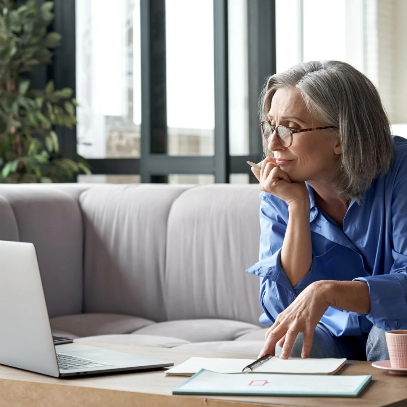 A woman with gray hair and glasses, wearing a blue shirt, sitting on a sofa, looking at a laptop, with a notebook and a pen in her hand, in a bright room with large windows.
