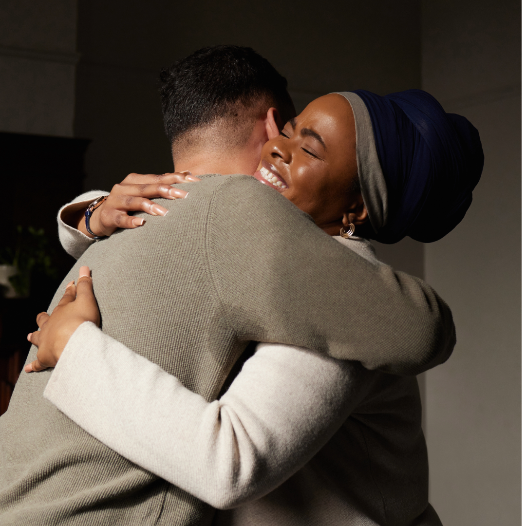 Two women hugging warmly, one with a headwrap, in an indoor setting.