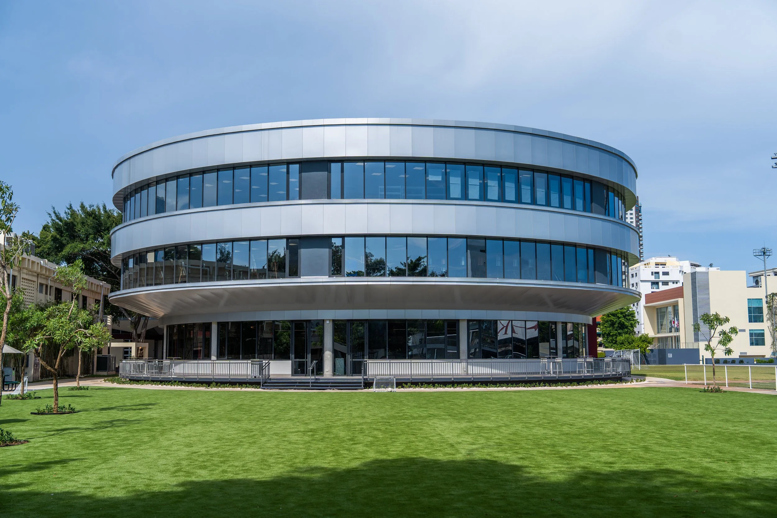Perspective rendering of the Carol Morgan School Arts & Dining Center designed by Arcoplan, showcasing a three-story circular building with expansive glass windows, curved balconies, and modern architectural lines, set against landscaped paths and at