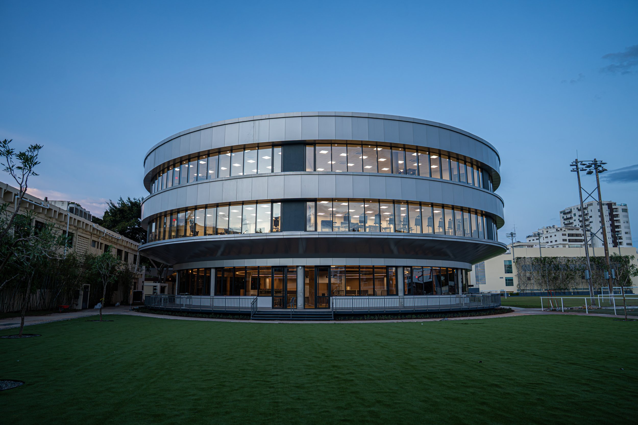 Exterior view of the Carol Morgan School Arts & Dining Center, a striking circular three-story building with wraparound terraces and expansive glass windows, set beside athletic fields and illuminated by stadium lighting in Santo Domingo.
