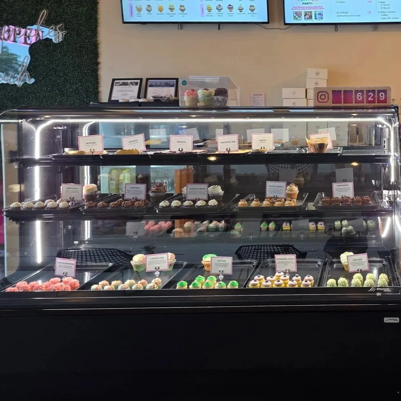 Display case filled with various cupcakes and desserts at a bakery, with digital menu screens and framed awards above.
