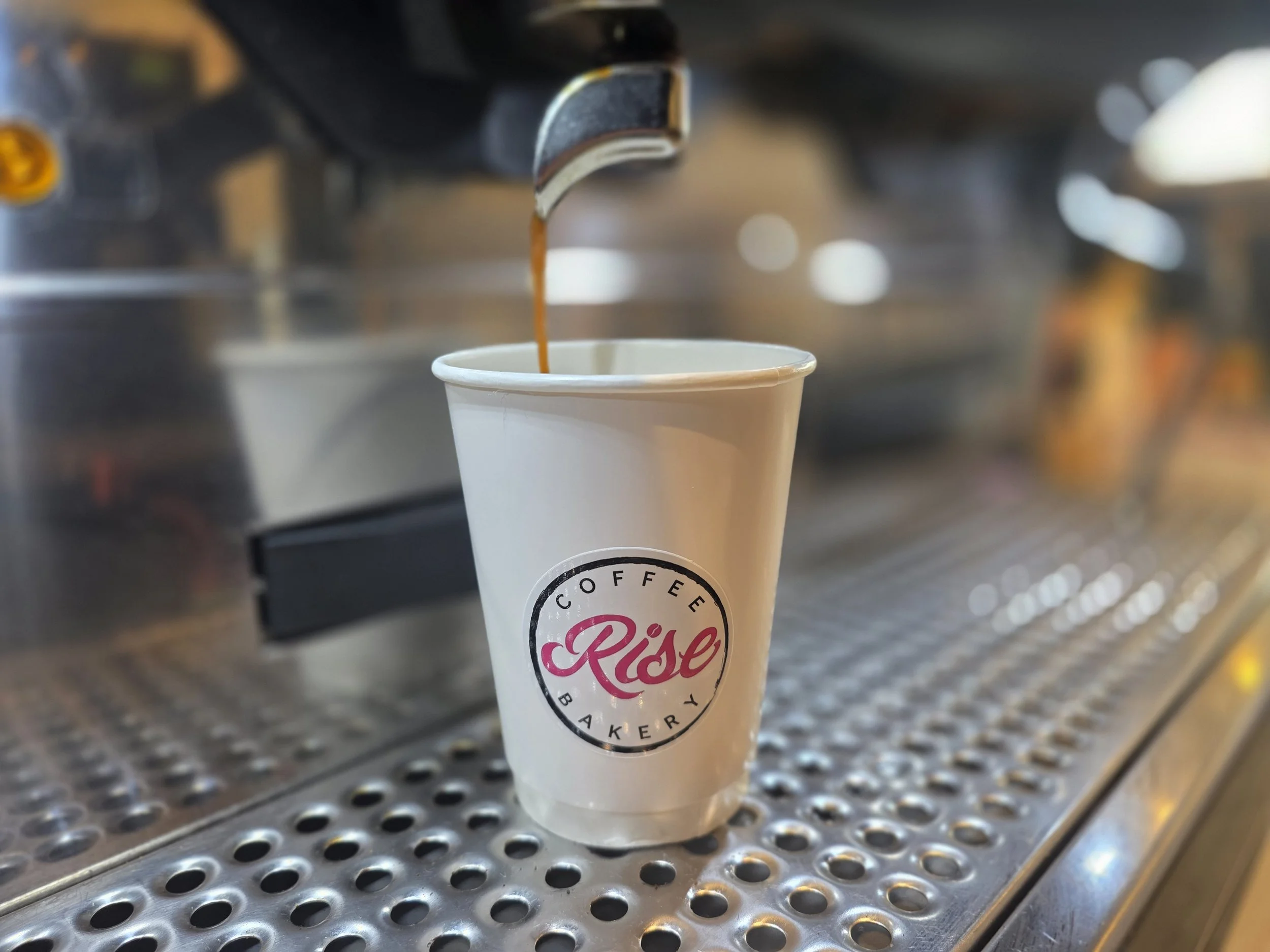 A coffee machine dispensing dark coffee into a white paper cup with a "Rise" logo on it, placed on a metal drip tray.