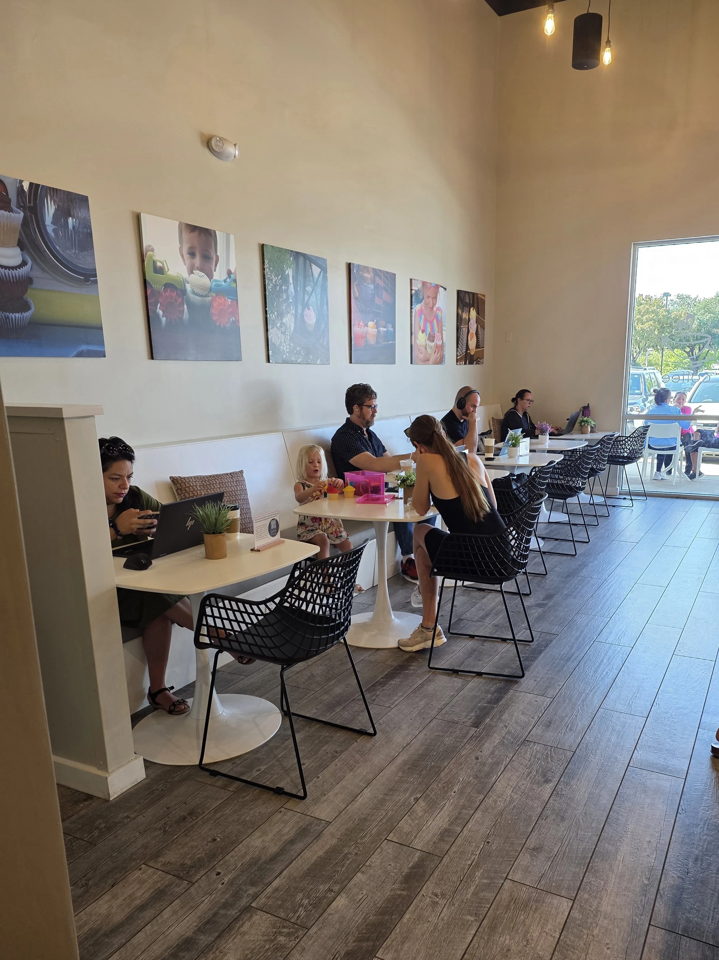 Interior of a coffee shop with customers seated at tables, some working on laptops, one person drinking, and a child playing with toys. Wall art features colorful photos of children and cupcakes, with large windows letting in natural light.