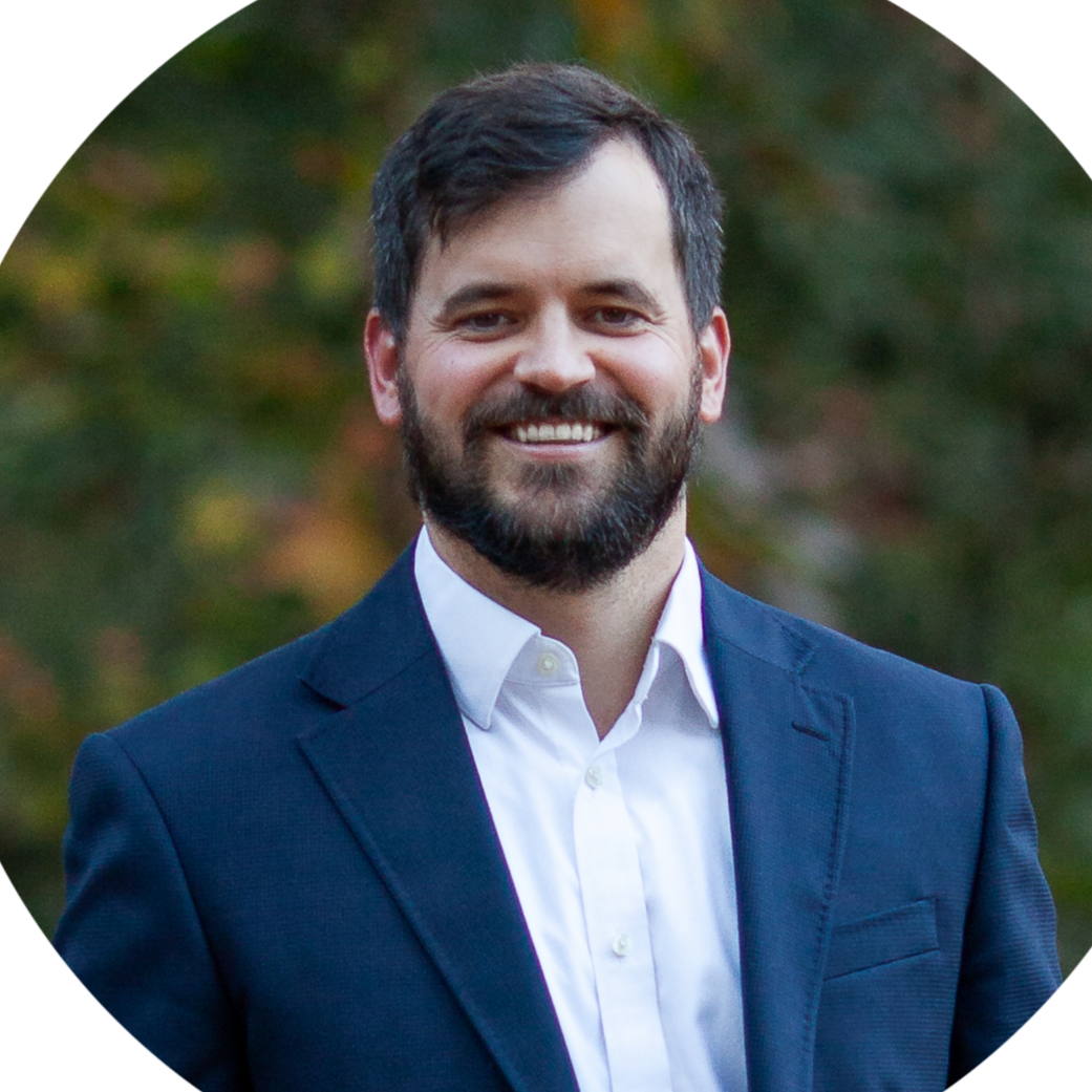 Professional headshot of a smiling man in a suit with a modern office building in the background.