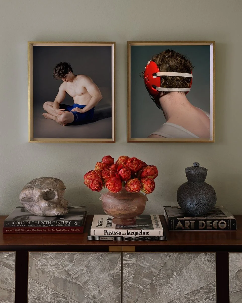A wooden console table with decorative books, a skull, and a vase of red flowers against a light green wall. Two framed photographs are hanging above; one shows a shirtless man sitting cross-legged, the other shows a person wearing a red helmet looking at the wall.