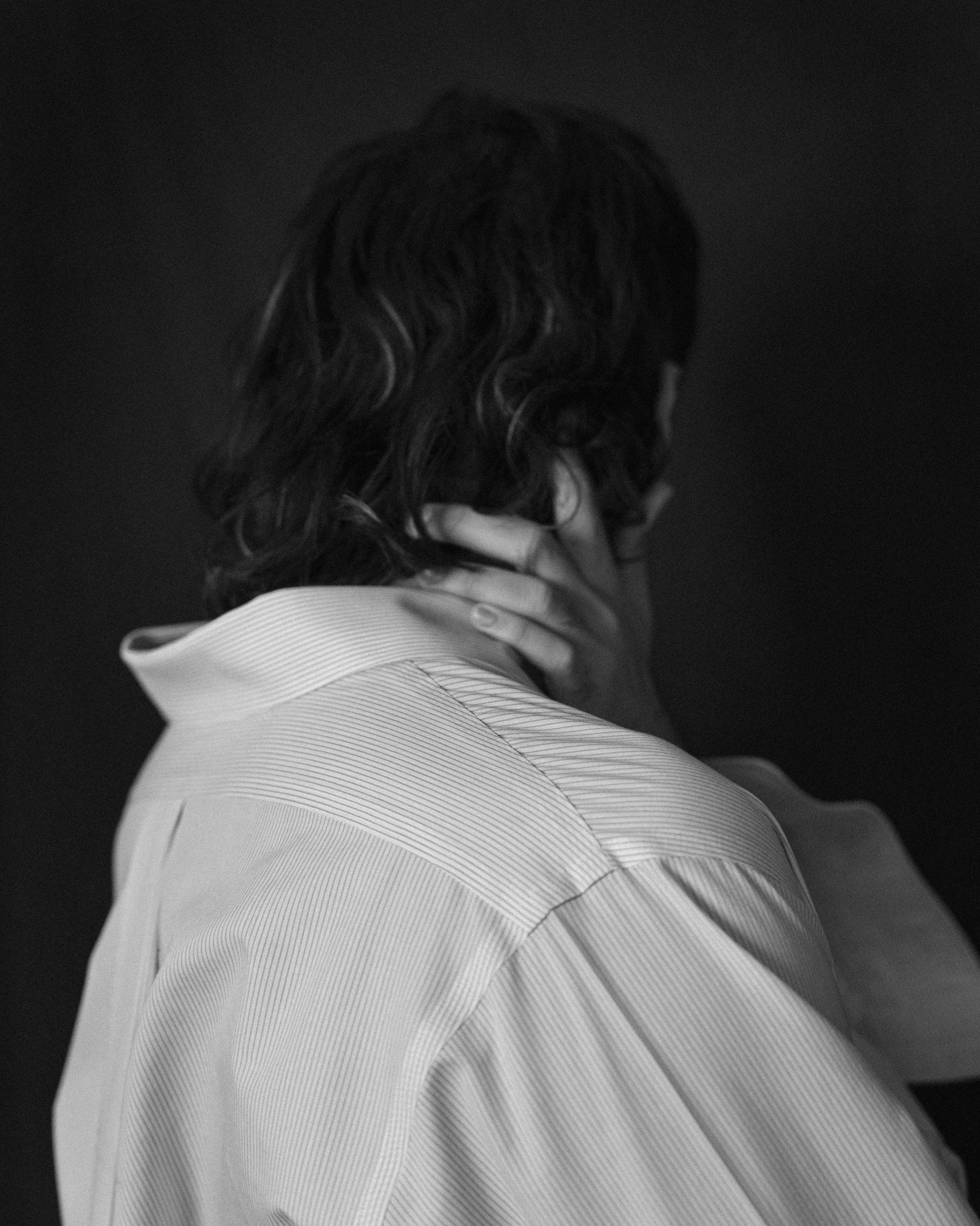 A person with curly hair wearing a light-colored striped shirt, touching their neck with one hand, against a dark background.