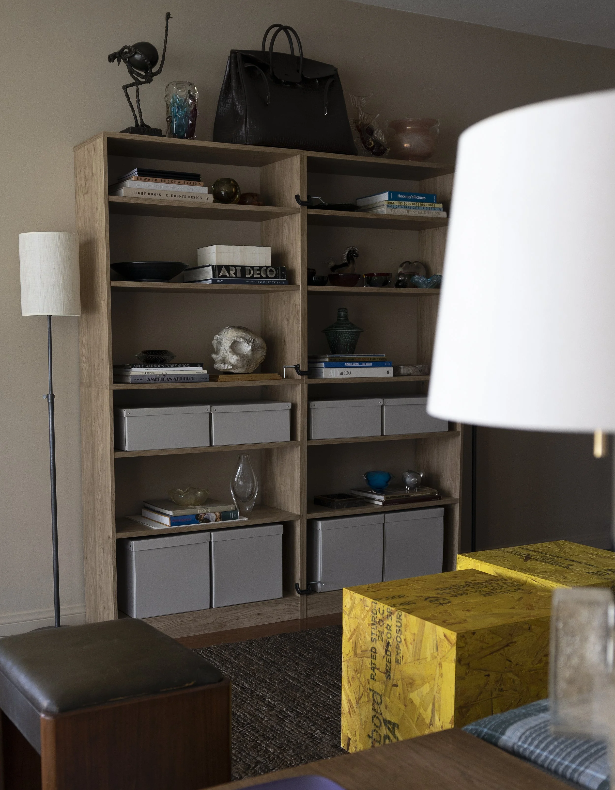 A living room with a tall wooden shelf filled with books, decorative objects, and storage boxes. A beige floor lamp stands to the left, and yellow boxes are on a table in the foreground. Part of a couch and a dark ottoman are also visible.