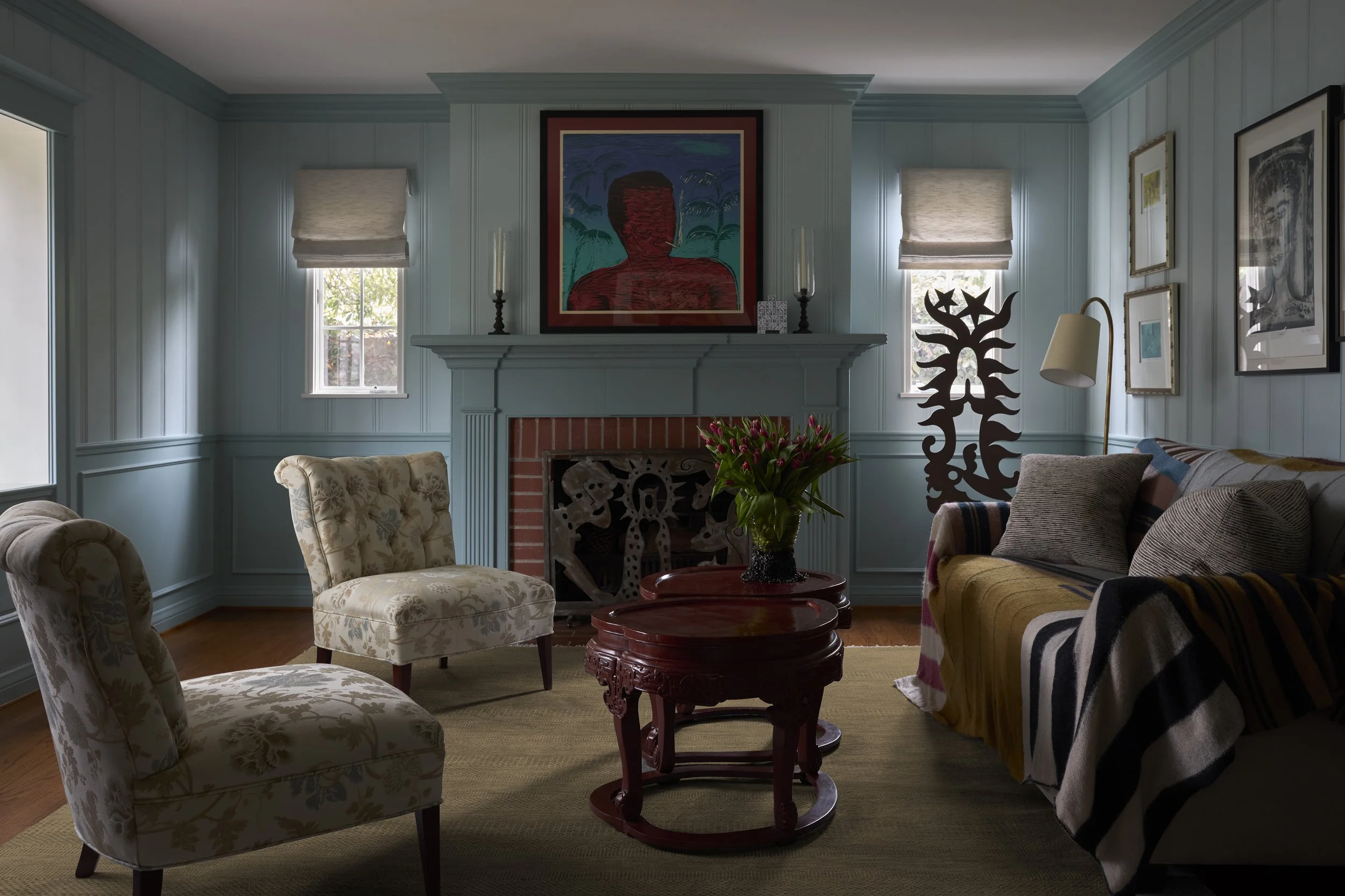 Living room with blue paneled walls, a brick fireplace with artwork above it, floral armchairs, a sofa with pillows, a wooden coffee table with a vase of pink tulips, and various framed pictures on the walls.